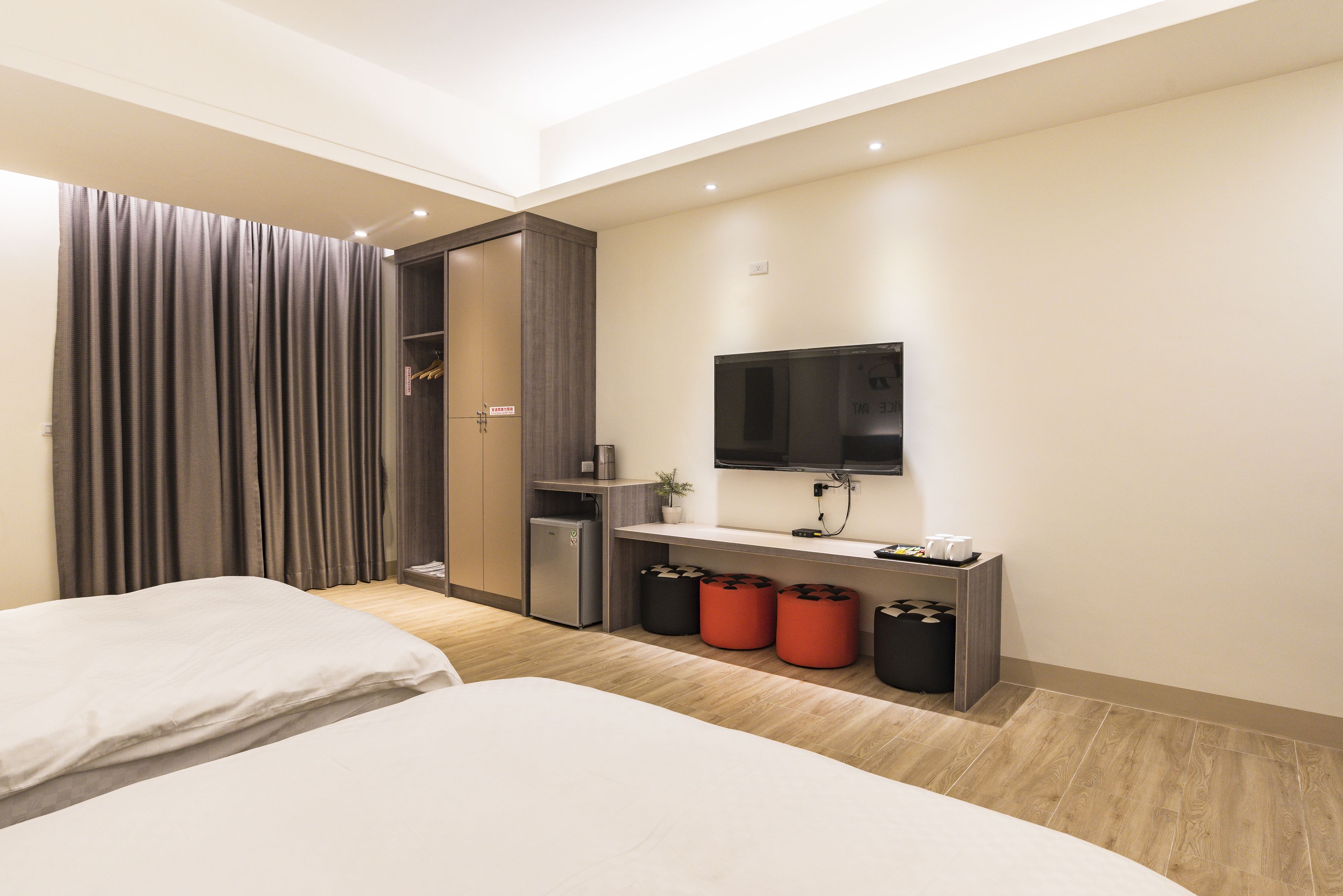 Quarto de hotel moderno com duas camas, uma TV, uma escrivaninha com cafeteira e uma pequena cozinha equipada.