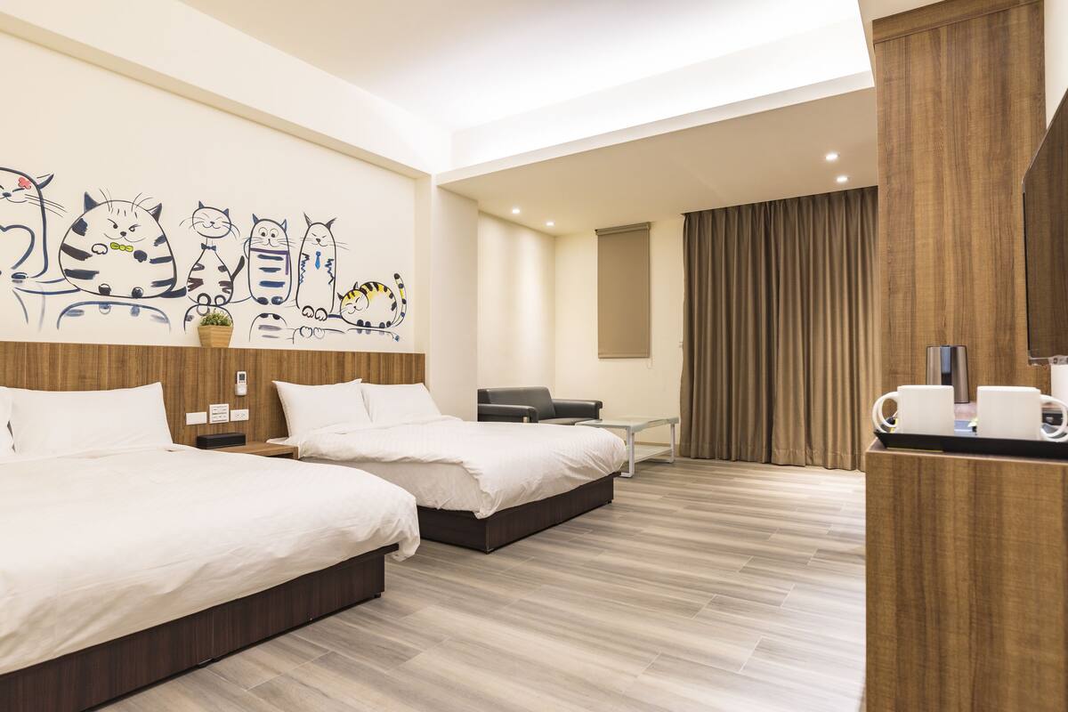 A hotel room with two beds, a desk, a chair, a wall with cat illustrations, and a wooden floor.