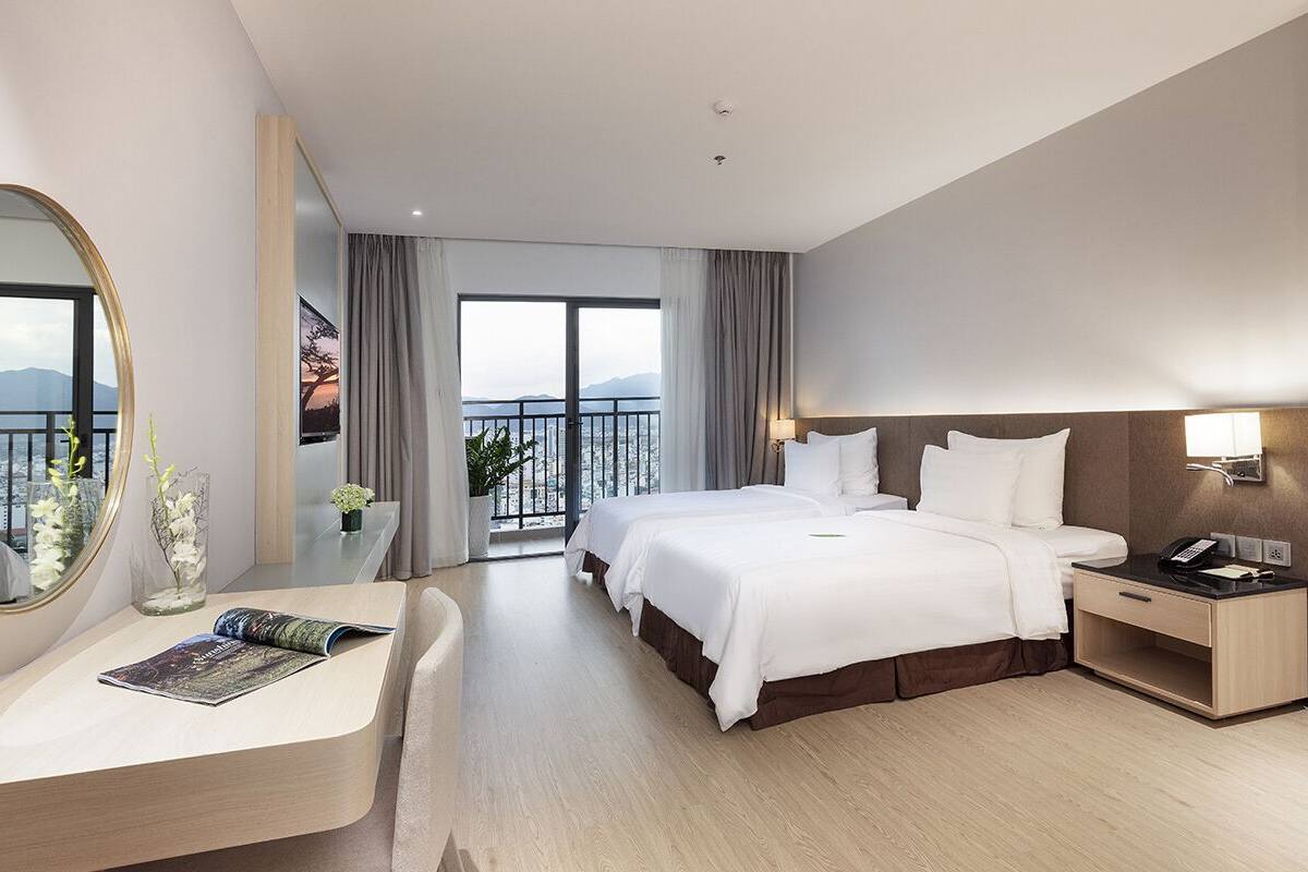 A modern hotel room with a large bed, a desk, a chair, a bedside table, and a view of the sea through large windows.