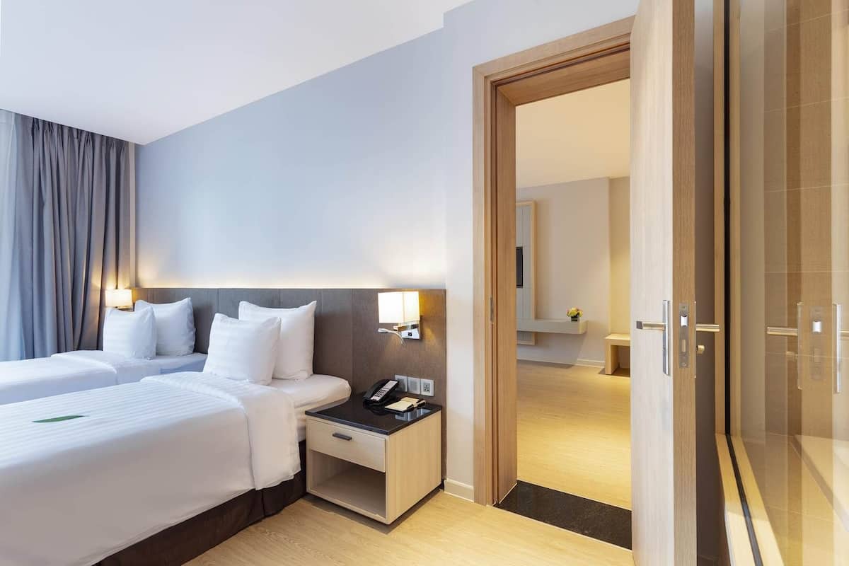 A hotel room with two beds, a bedside table with a phone and notepad, a nightstand with a lamp, and a wooden door leading to another room.