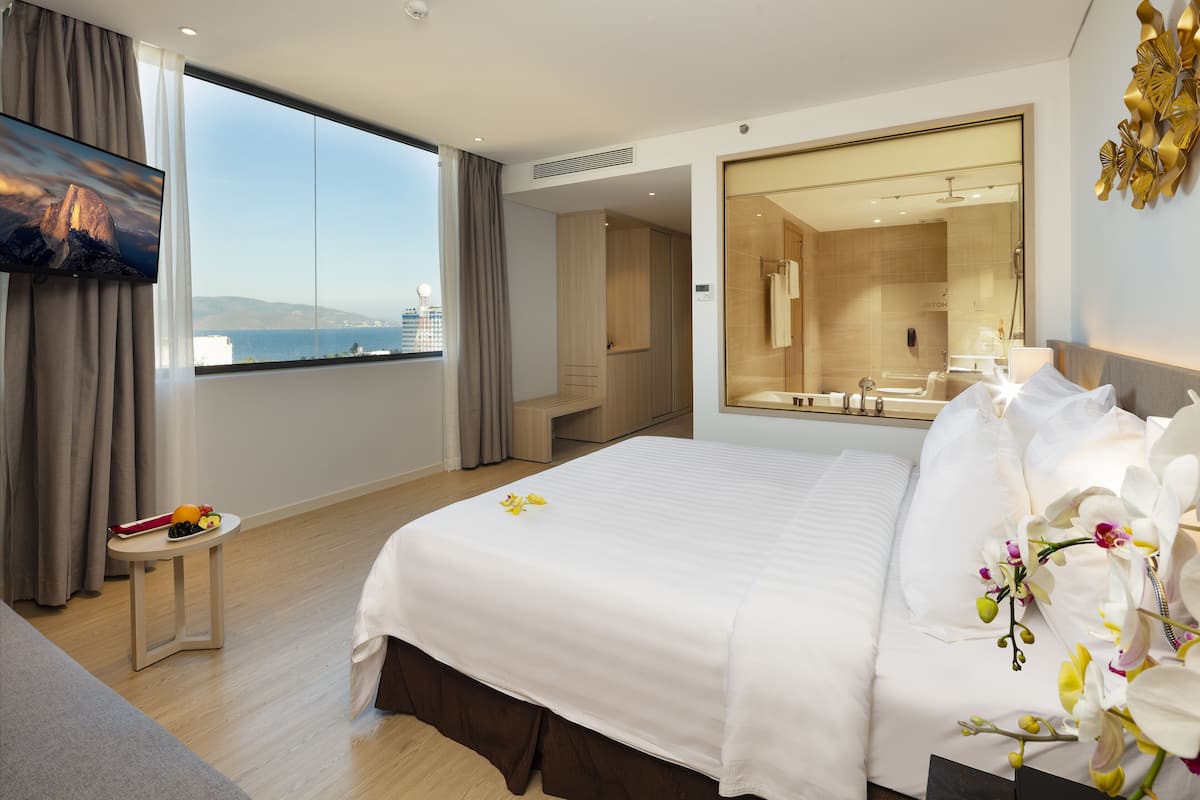 A hotel room with a large bed, a TV, a small table with fruit, a view of the sea, and a bathroom with a mirror and shower.