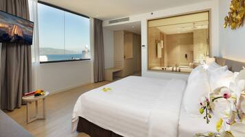 A hotel room with a large bed, a TV, a small table with fruit, a view of the sea, and a bathroom with a mirror and shower.