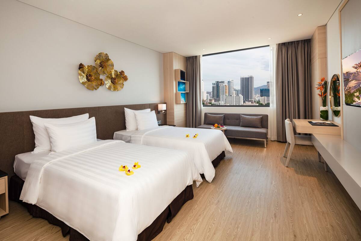 A hotel room with two beds, a desk, a sofa, a city view, and decorative wall art.