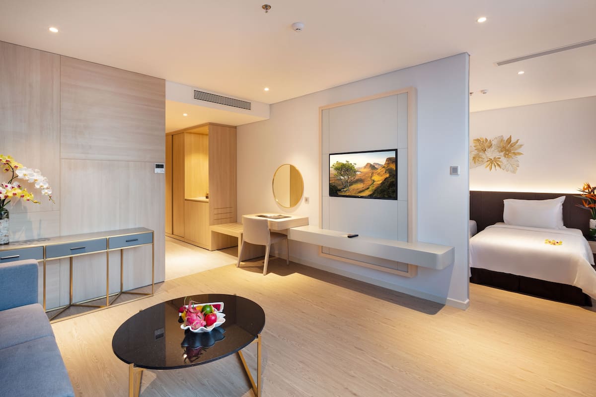 A modern hotel room with a bed, a desk, a chair, a small table with fruit, and a TV on the wall.