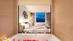 A modern hotel room with a large bathtub filled with rose petals, a bed with a city view, and a cozy seating area. - DQua Hotel (Nha Trang)