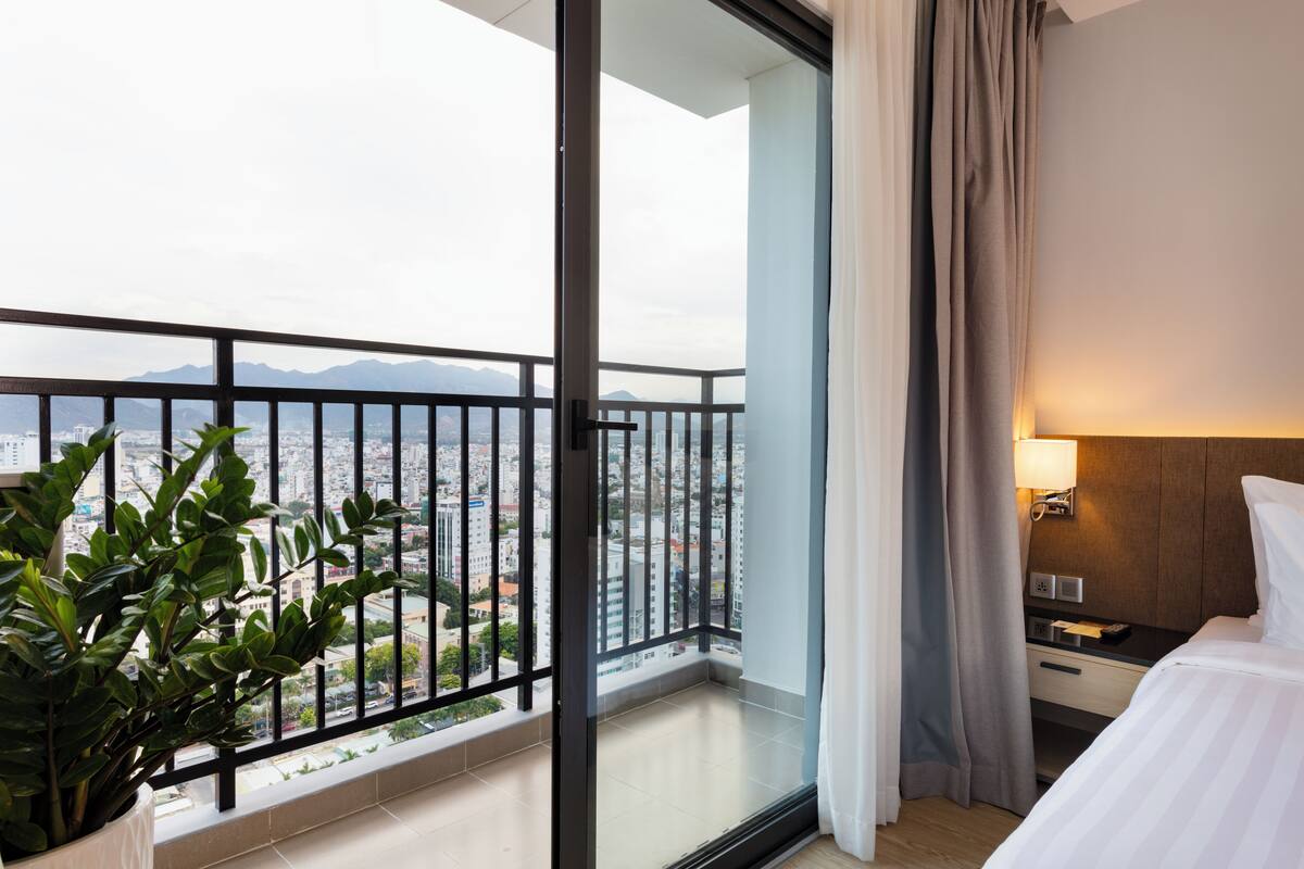A hotel room with a balcony, a bed, a nightstand, a lamp, and a potted plant.