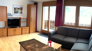 A modern living room with a sofa, a television, wooden cabinets, and a large window with a view of a balcony. - Apartment Azurit by Interhome (Saas-Fee)