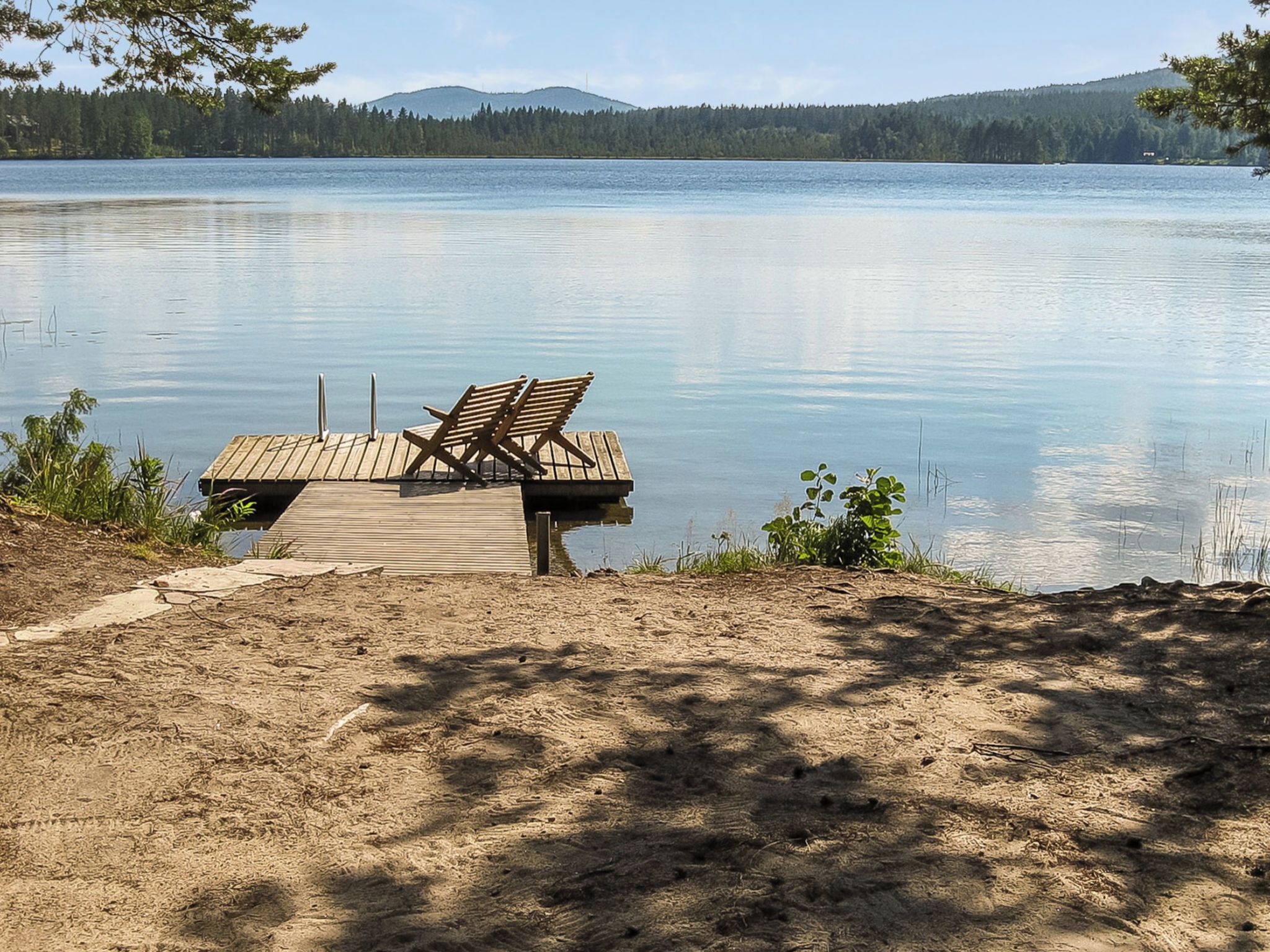 Eine Holzstele mit einer Bank erstreckt sich über einen ruhigen See, umgeben von Bäumen und einem klaren Himmel.