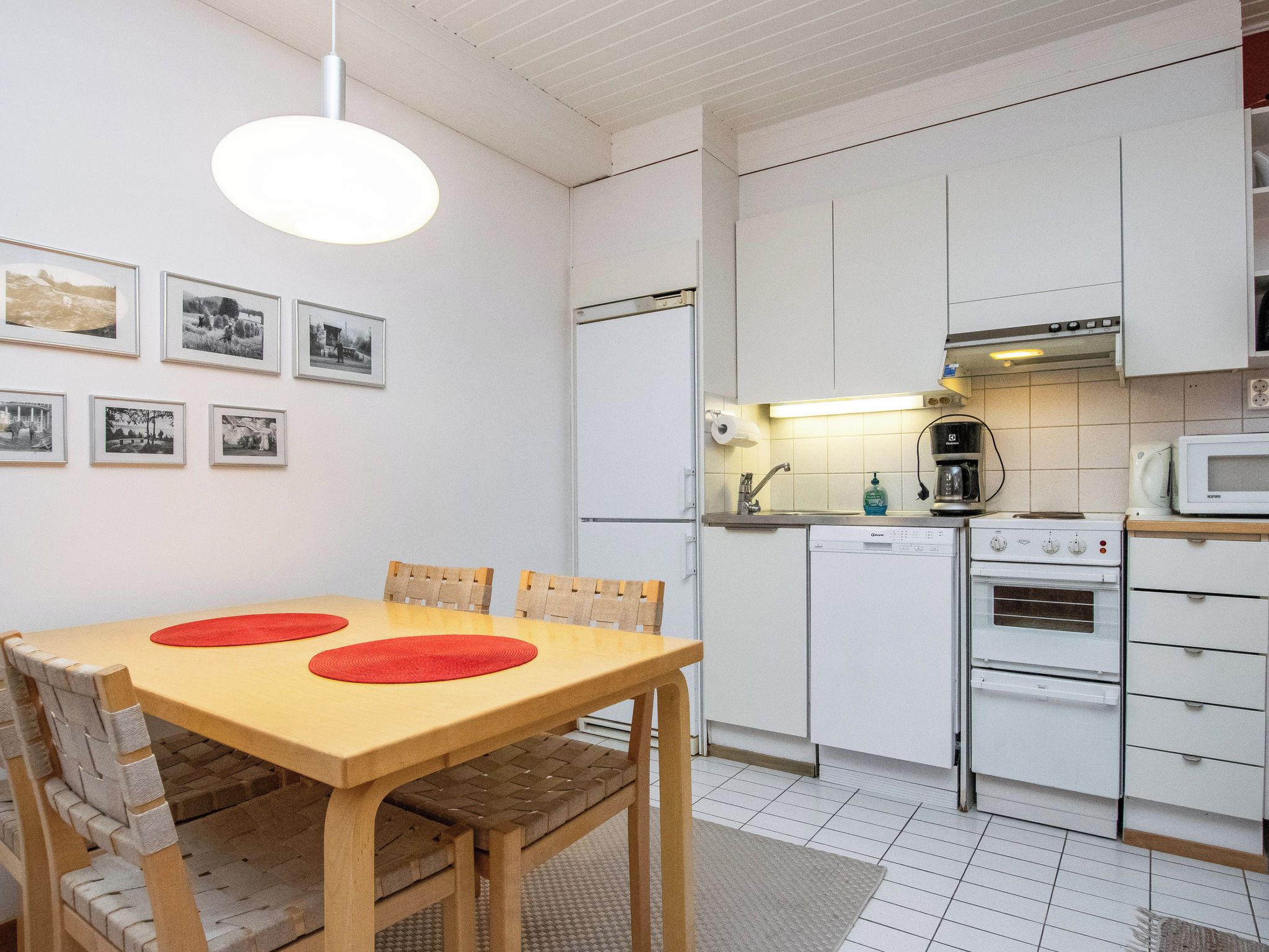 Une cuisine compacte avec des meubles blancs, une table à manger en bois agrémentée de sets de table rouges et un luminaire suspendu.