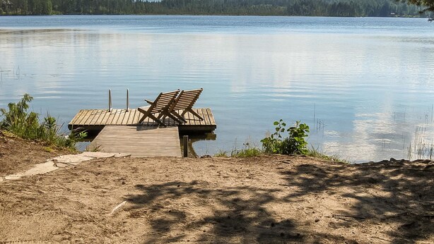 Un muelle de madera con dos sillas se adentra en un lago tranquilo, rodeado de un paisaje boscoso.