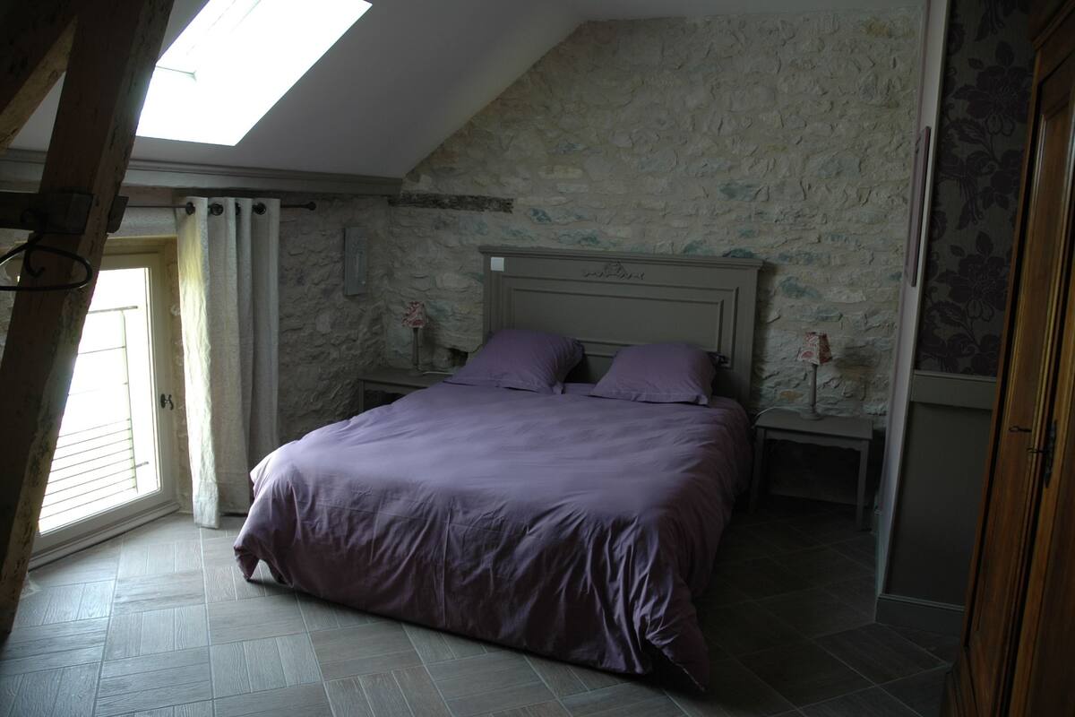 Un dormitorio con pared de piedra, cama con ropa de cama púrpura, mesita de noche y ventana con cortinas.