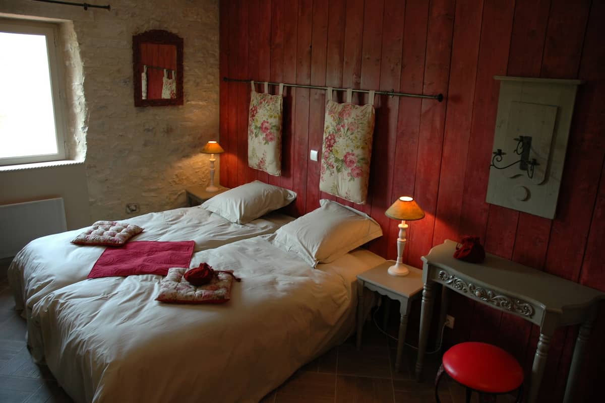 Un dormitorio con dos camas, una mesita pequeña, un taburete rojo y una lámpara fijada a la pared.
