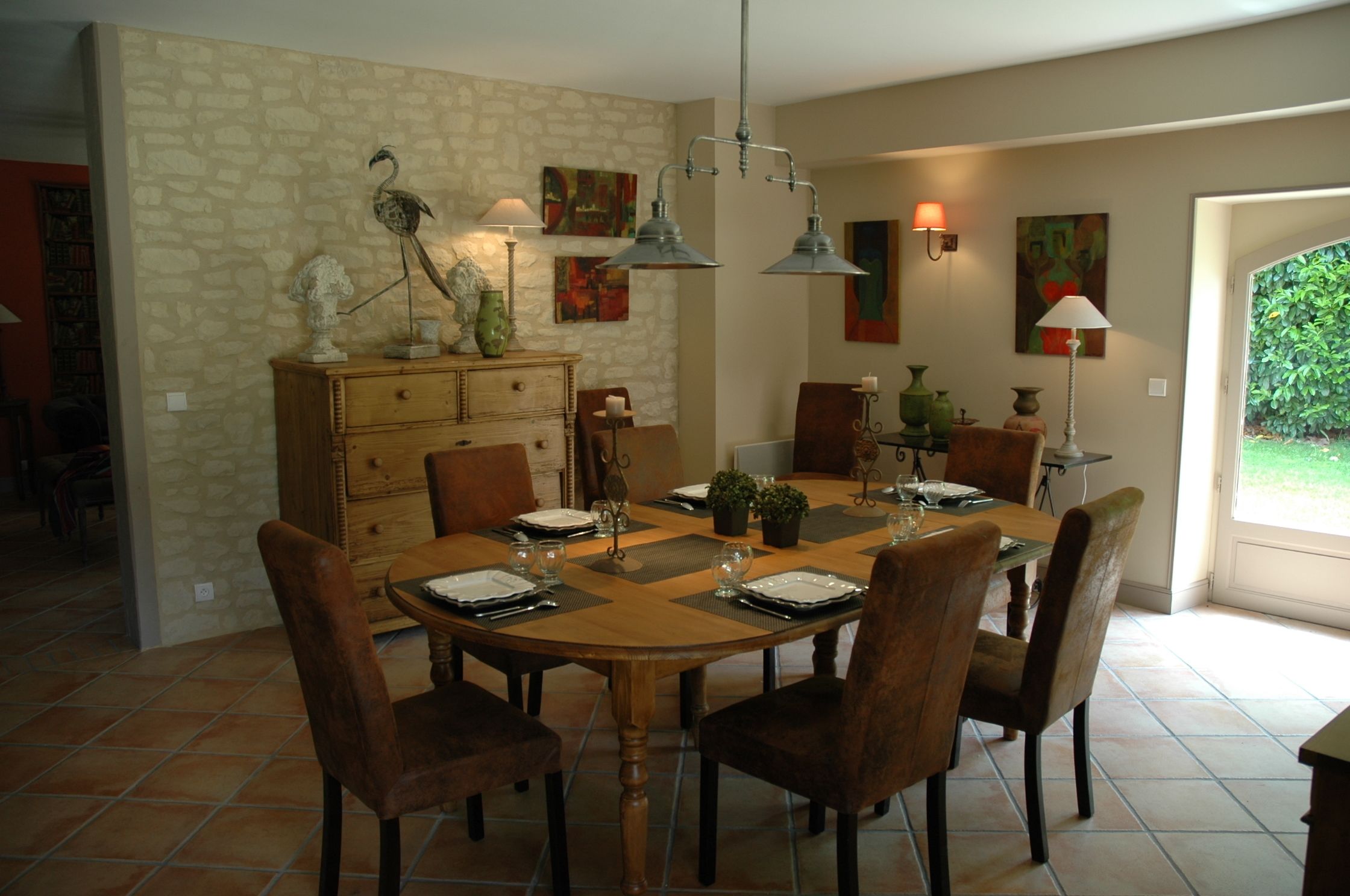 A dining area with a round wooden table set for six, surrounded by six chairs. There is a stone wall with a wooden cabinet and decorative items on top. Two wall-mounted lamps provide lighting.