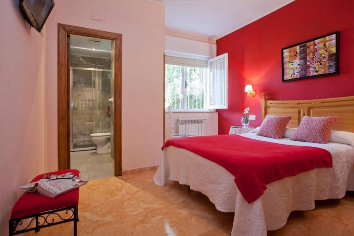 A bedroom with a bed, a red wall, a window, and a door leading to a bathroom.