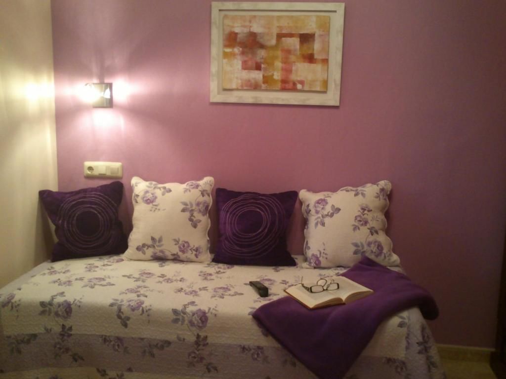 Quarto com cama, almofadas roxas, uma obra de arte emoldurada e uma mesa de cabeceira com um livro e óculos.