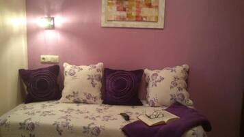 Quarto com cama, almofadas roxas, uma obra de arte emoldurada e uma mesa de cabeceira com um livro e óculos.