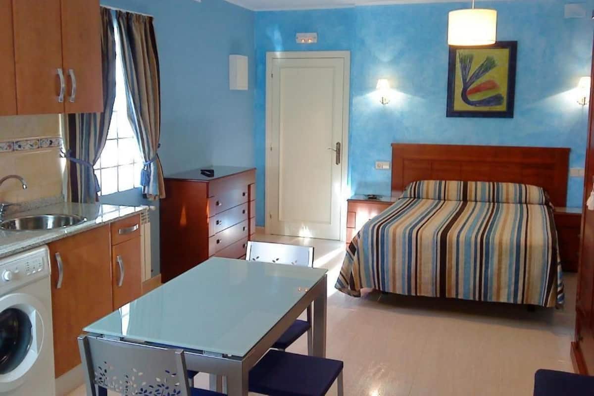 A compact hotel room with a bed, a small table, a chair, a washing machine, and a kitchenette.