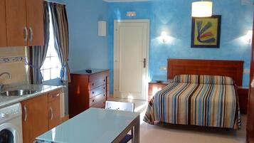 Habitación de hotel compacta con cama, una mesita, una silla, lavadora y kitchenette.