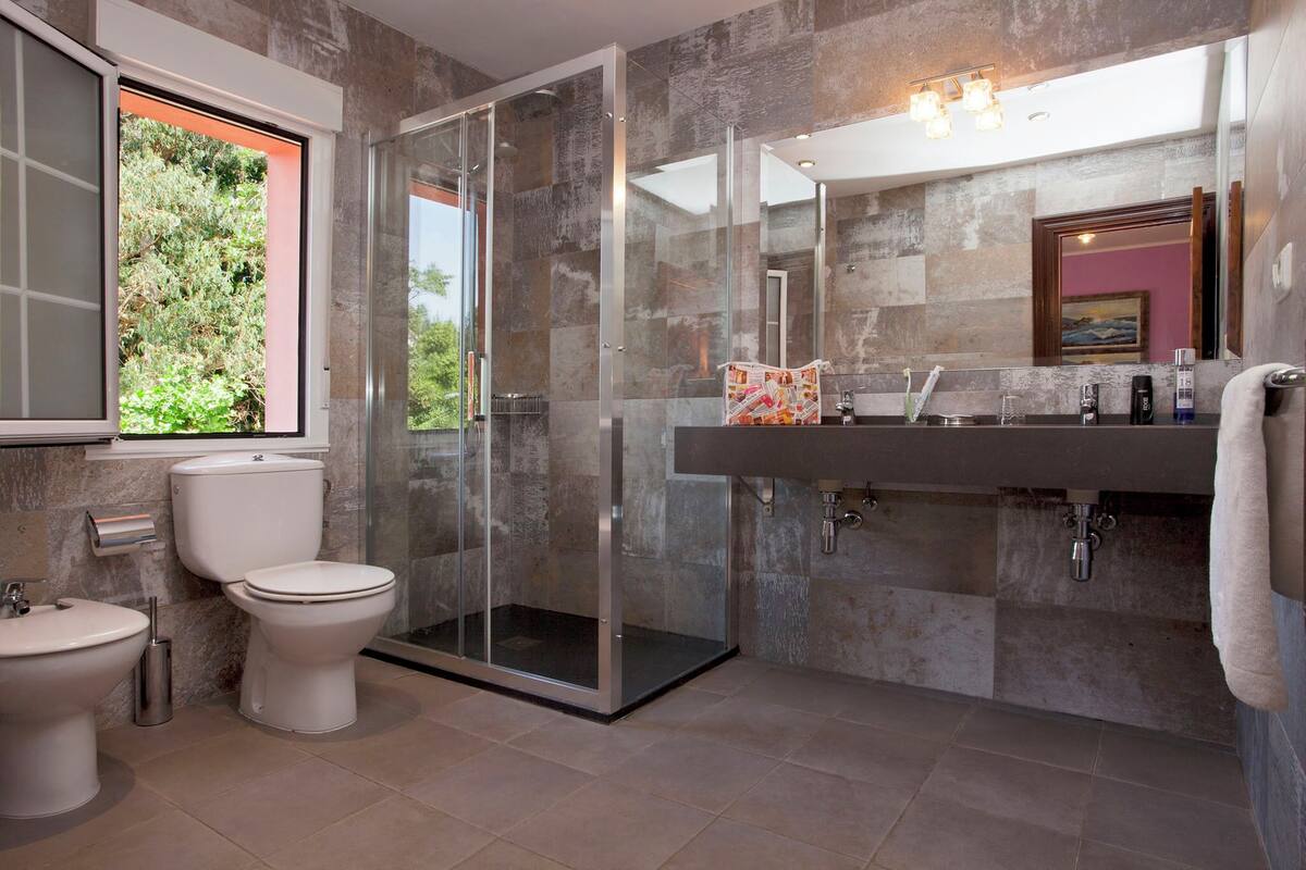 A modern bathroom with a glass-enclosed shower, a large mirror, and a window with a view of greenery.