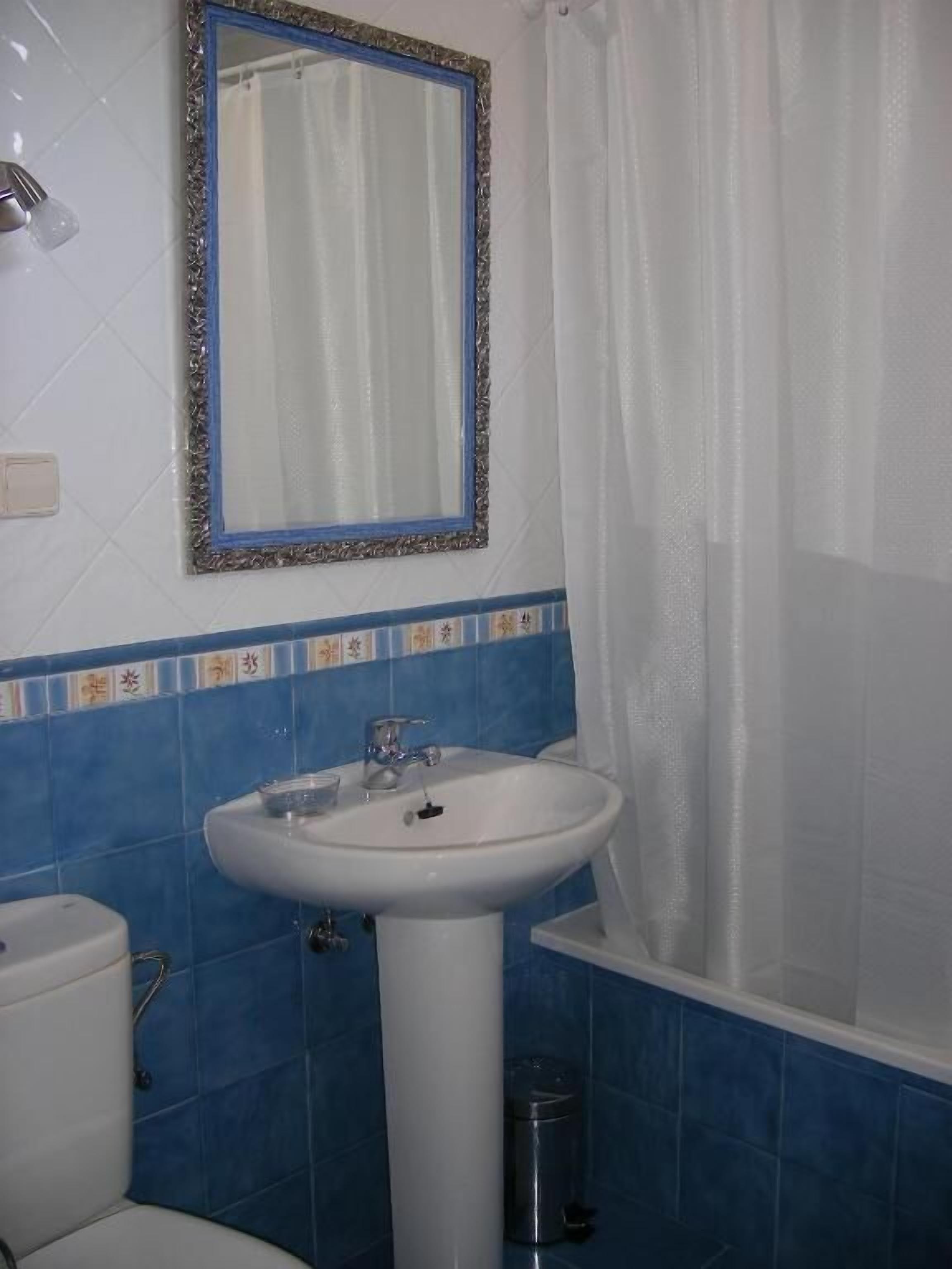 Un baño con un lavabo blanco, paredes con azulejos azules, un inodoro blanco y una bañera con cortina de ducha blanca.