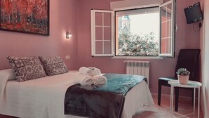 A bedroom with a bed, pillows, a window with a view, a radiator, a chair, and a small table with a plant. - Hotel Alvaro (Cudillero)