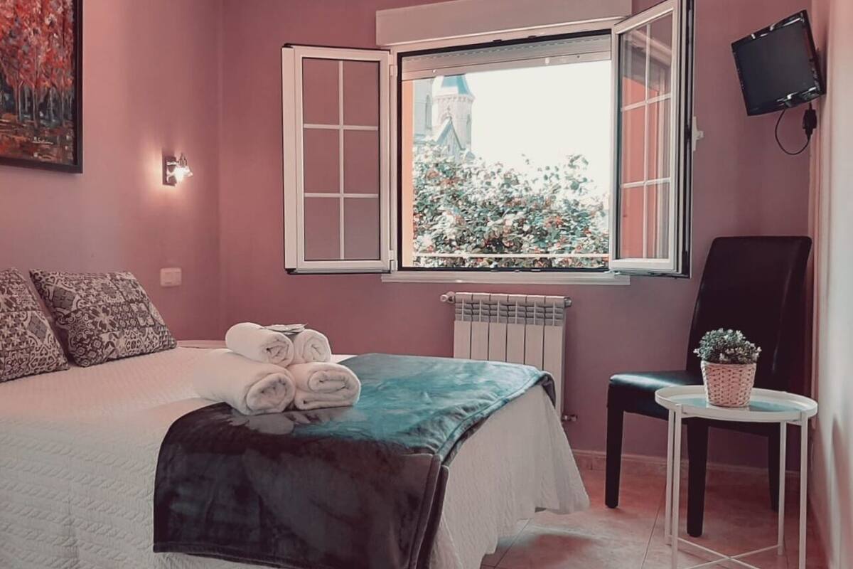 A bedroom with a bed, pillows, a window with a view, a radiator, a chair, and a small table with a plant.