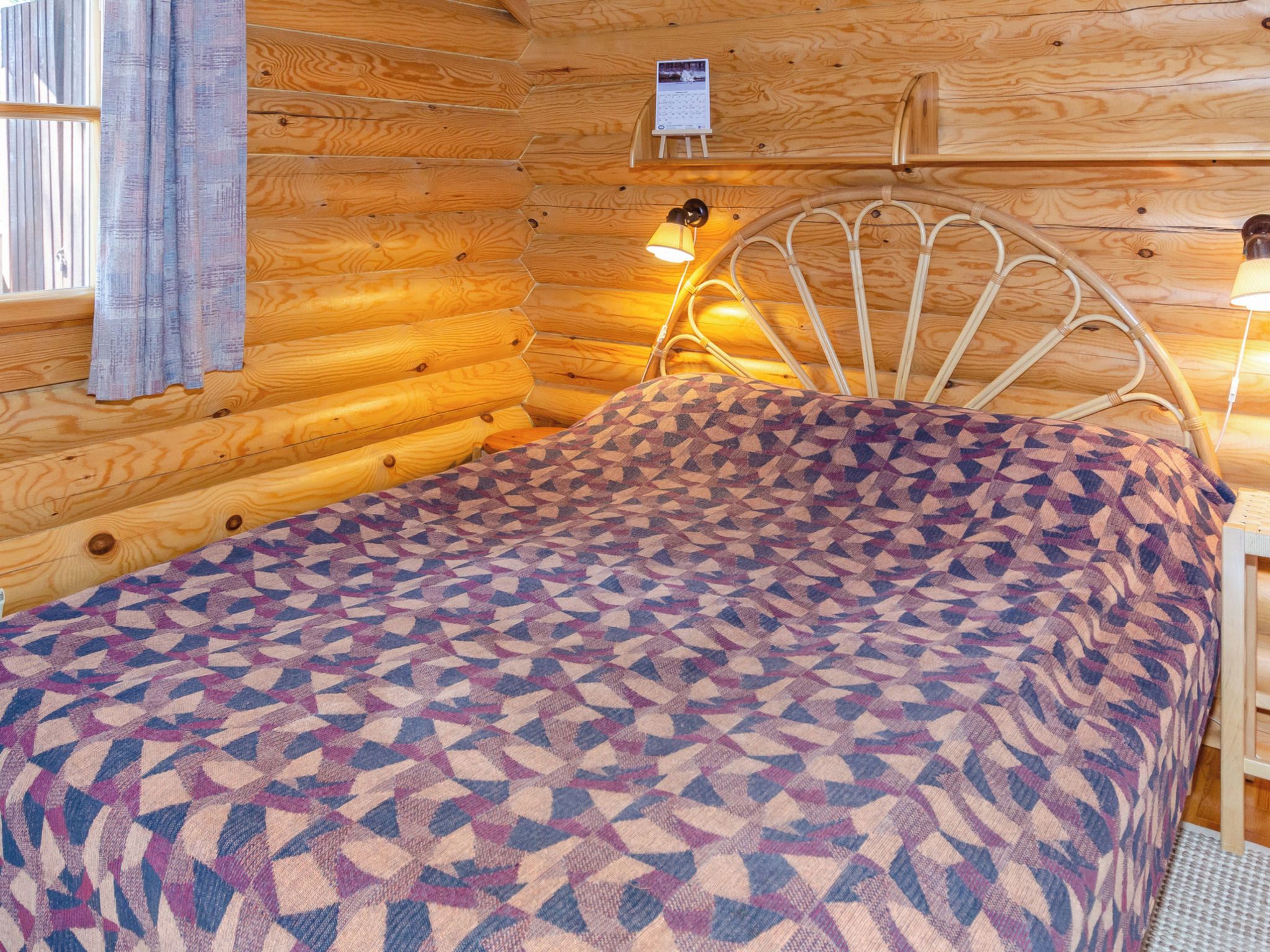 A wooden cabin interior with a bed, a wall calendar, and a window with curtains.