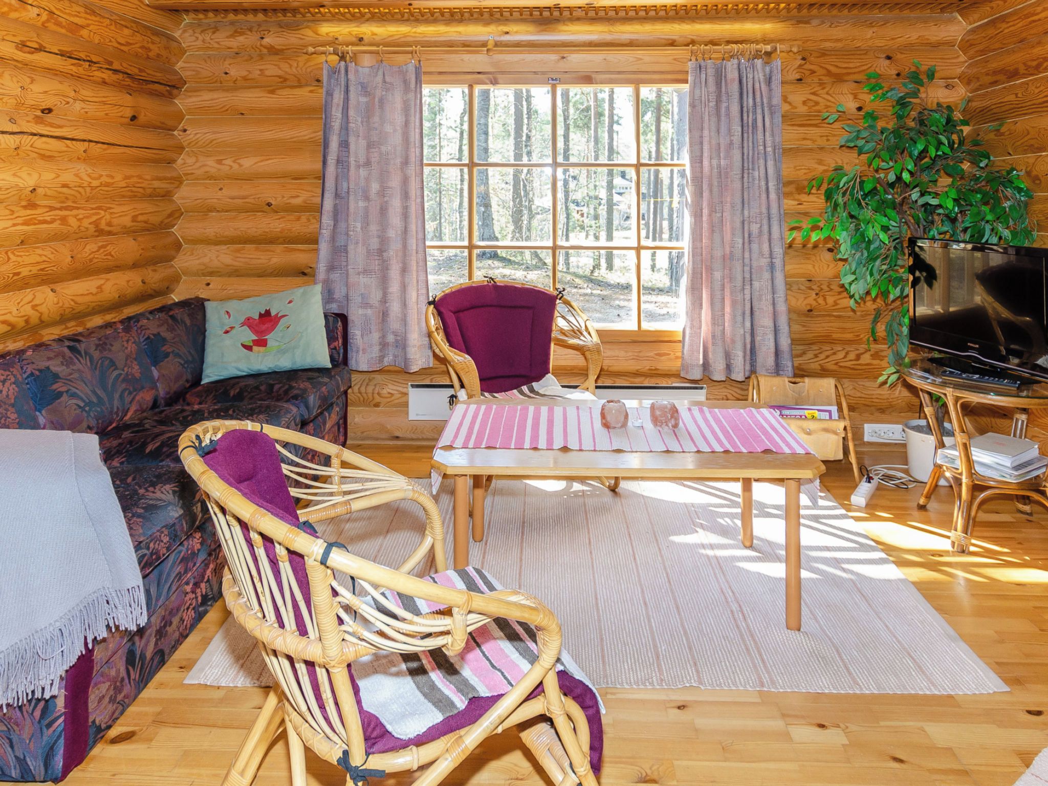 A log cabin interior with a sofa, wicker chairs, a table, a television, and a window with curtains.