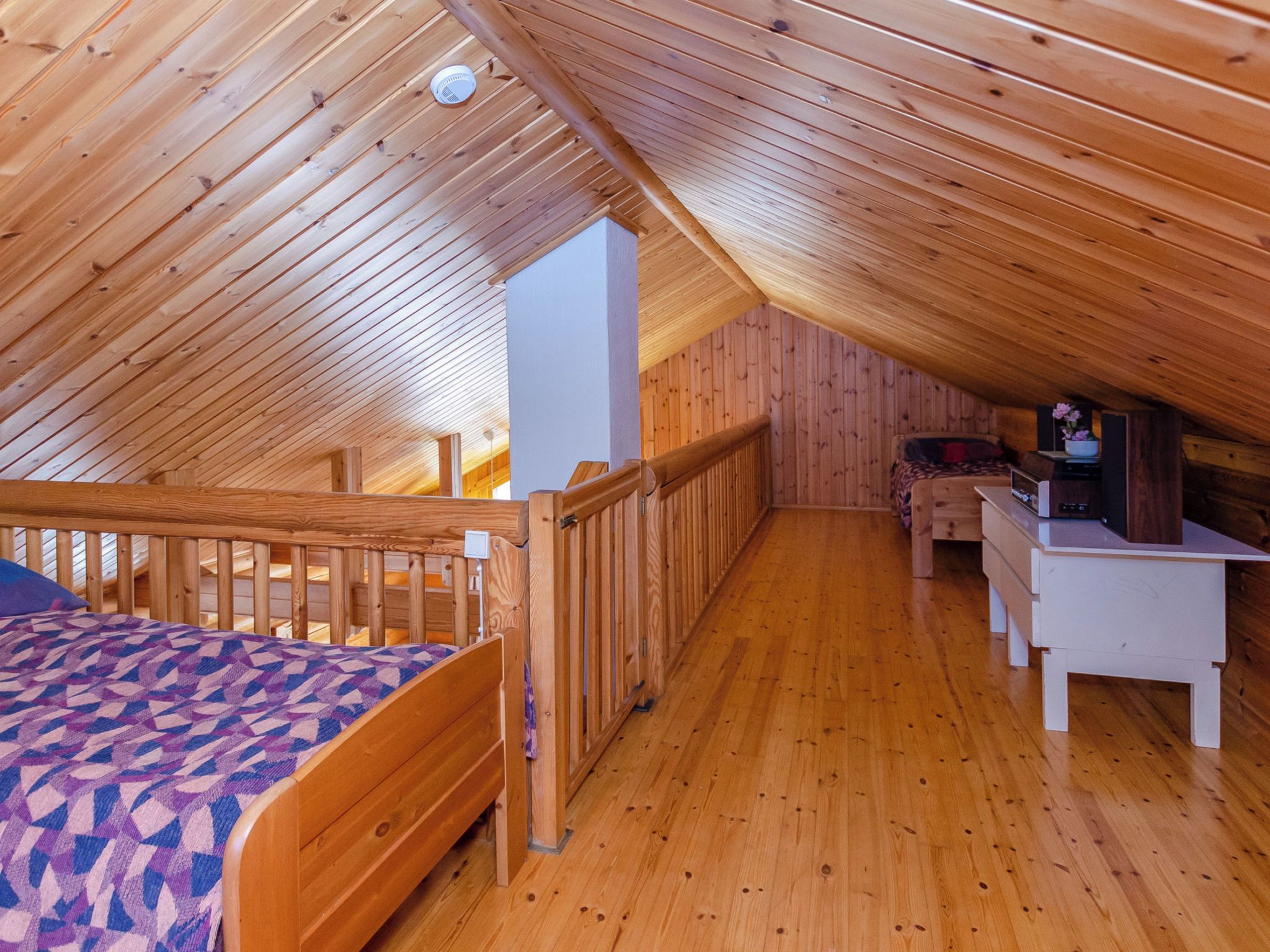 A wooden interior with a sloped ceiling, a bed with a patterned blanket, a desk with a computer, and a chair.