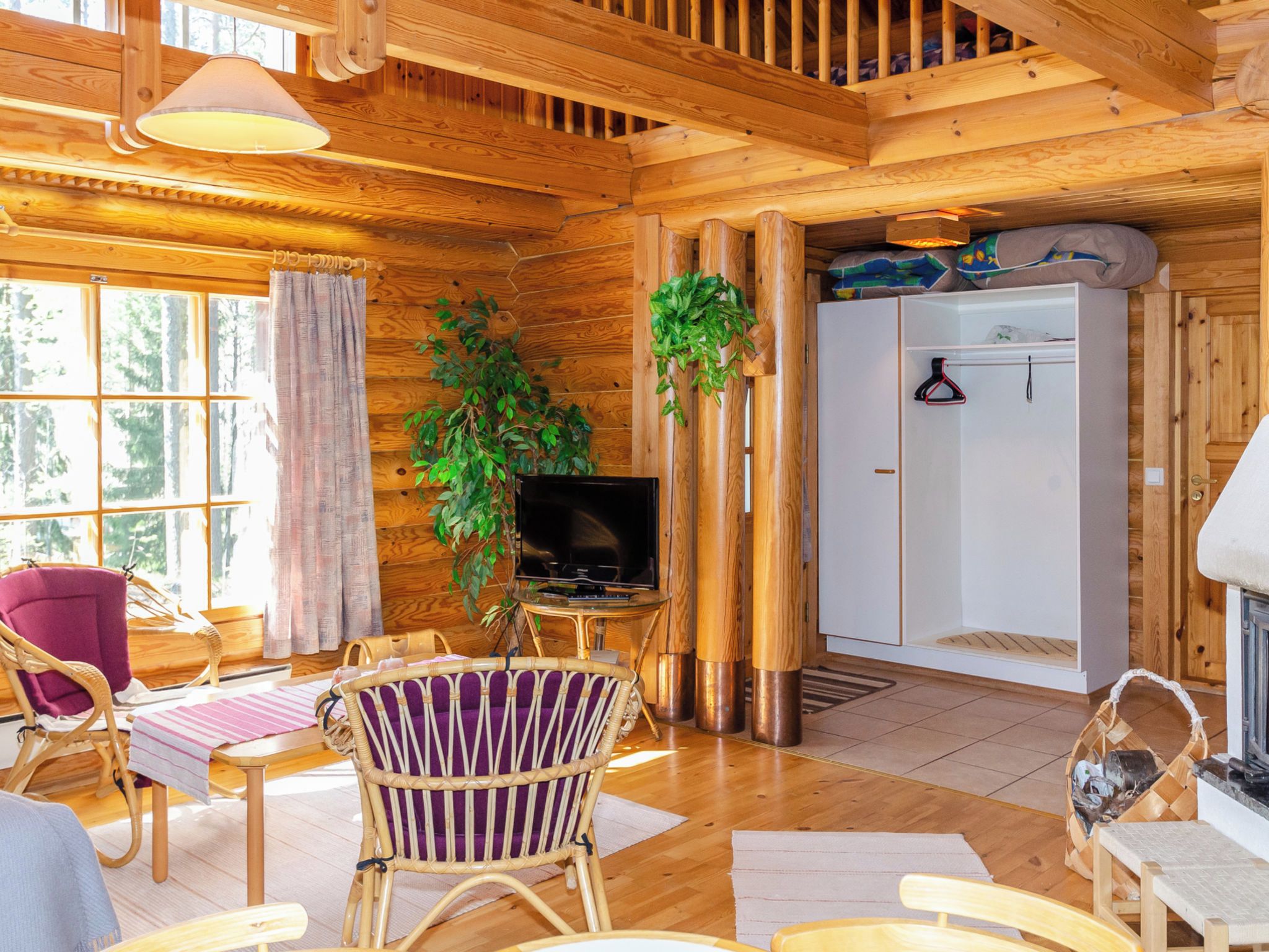 A wooden cabin interior with a television, wicker furniture, a fireplace, and a large window.