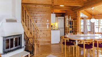 A wooden cabin interior with a fireplace, wooden staircase, kitchen area, dining table with chairs, and large windows.