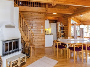 Interior de una cabaña de madera con chimenea, escalera de madera, zona de cocina, mesa de comedor con sillas y ventanas amplias.