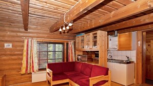 Un accogliente interno di una baita con un divano in legno, un tavolo da pranzo, un angolo cottura e un'ampia finestra con tende.