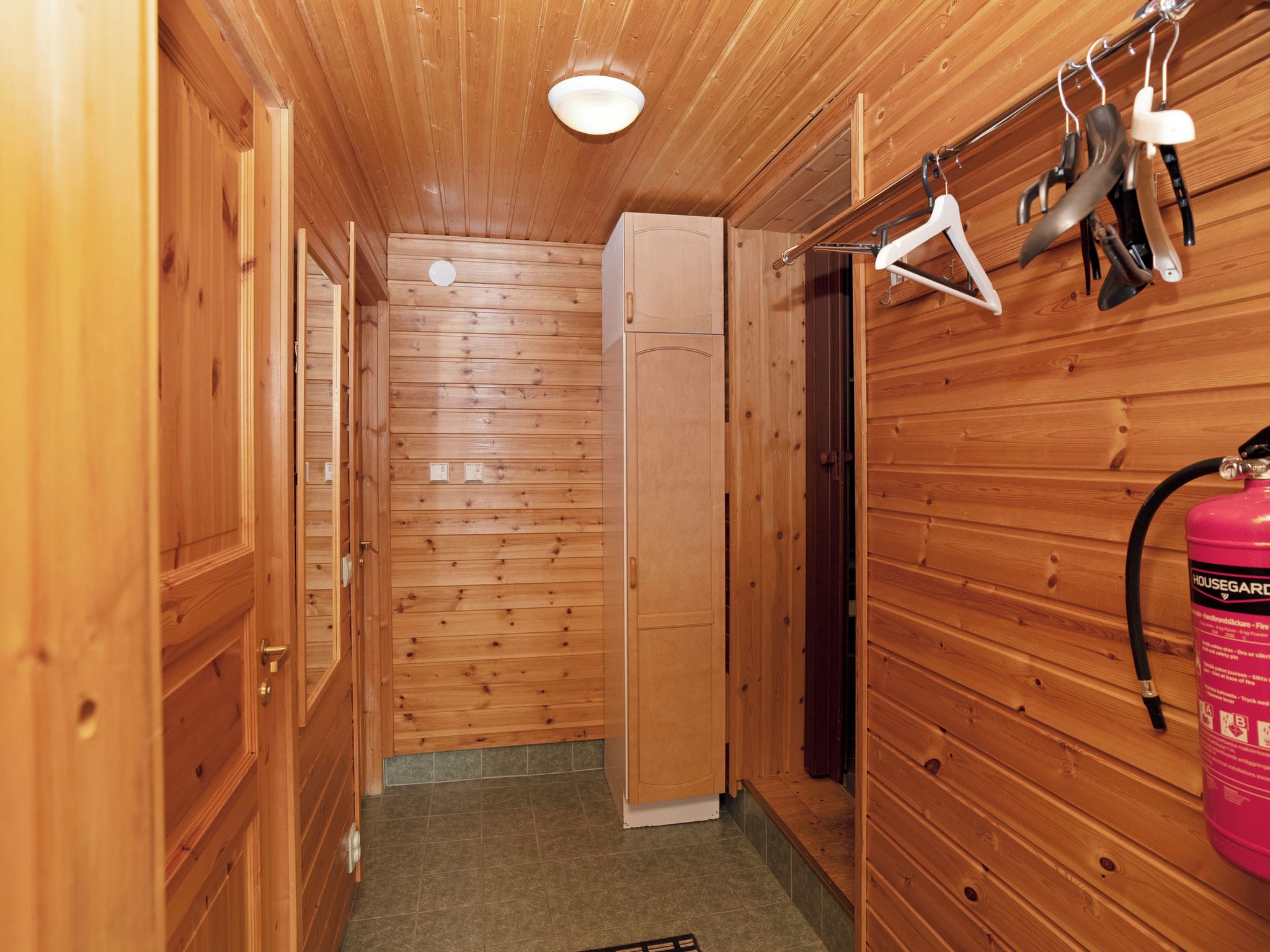 Un intérieur en bois avec un extincteur fixé au mur, une porte donnant sur une pièce et un luminaire au plafond.