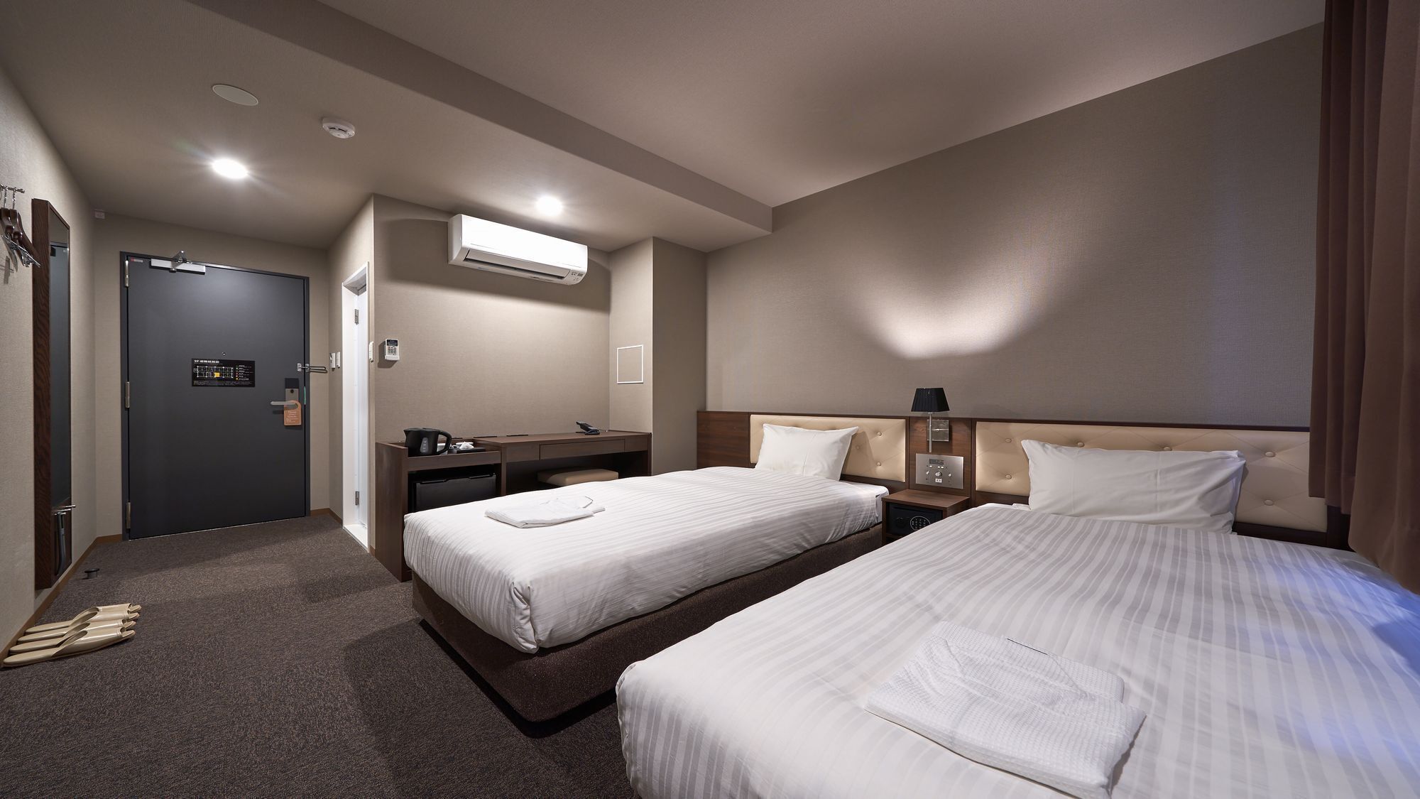 A hotel room with two beds, a desk, a chair, a wardrobe, and an air conditioning unit.