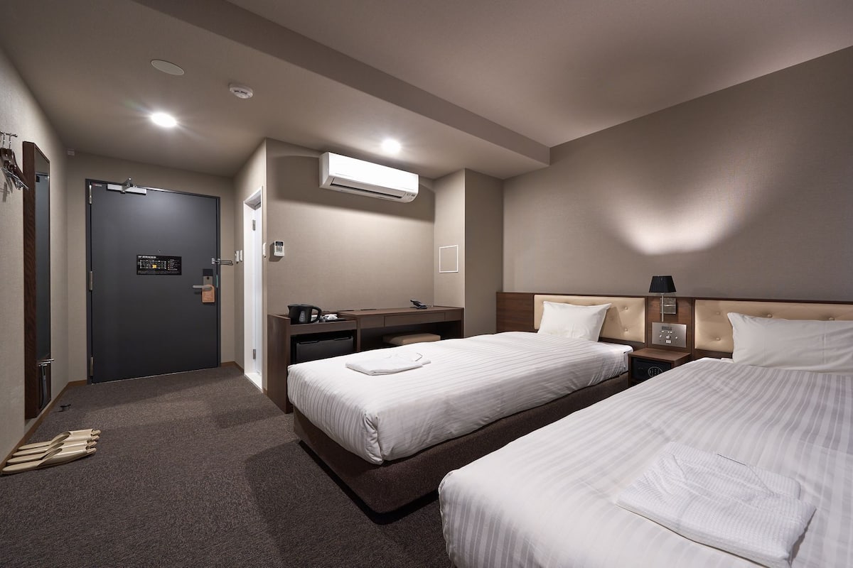 Ein Hotelzimmer mit zwei Betten, einem Schreibtisch, einem Stuhl, einem Kleiderschrank und einer Klimaanlage.