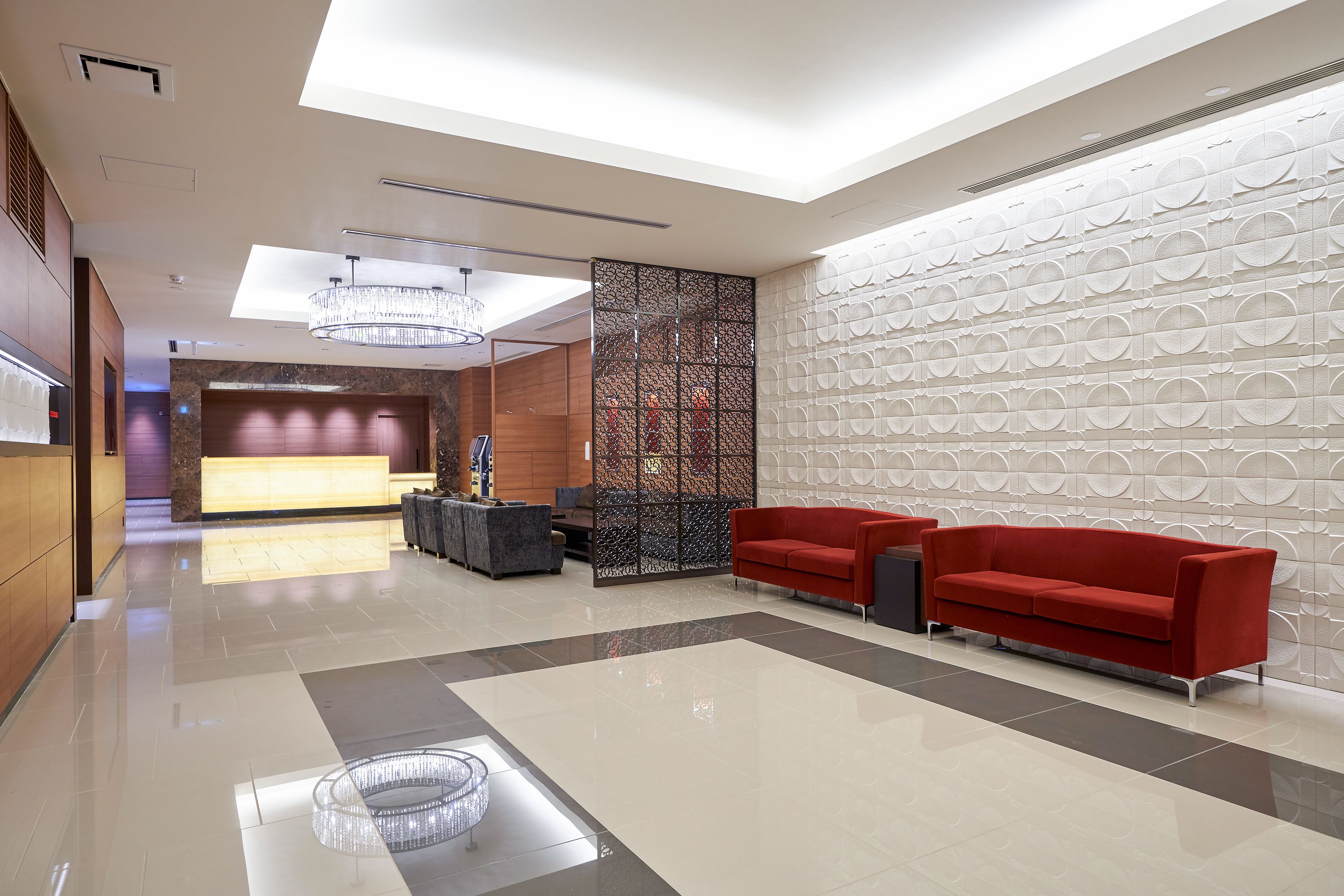 A modern lobby with a reception desk, red sofas, a decorative wall, and a chandelier.