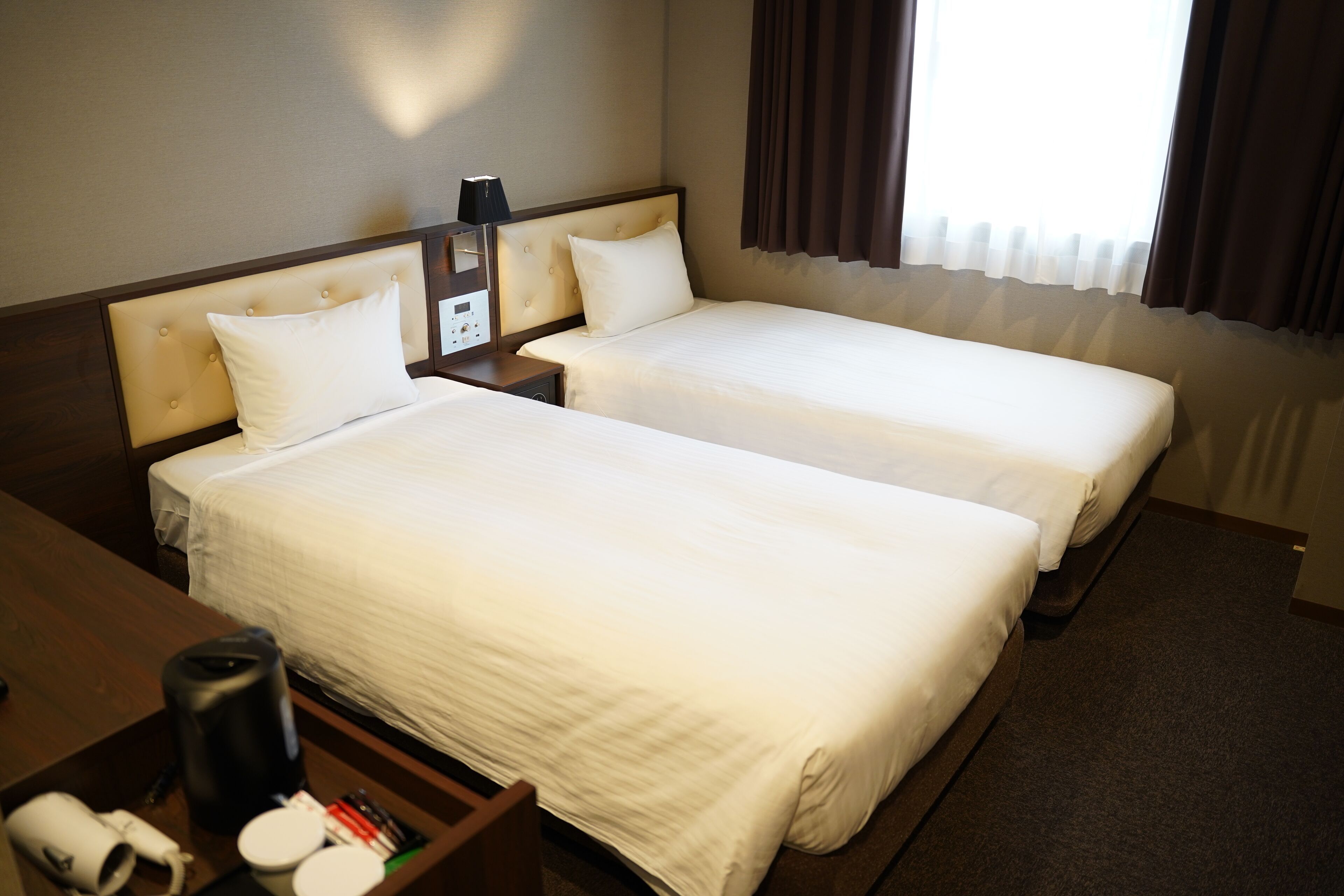 A hotel room with two single beds, a nightstand with a lamp, a coffee maker, and a small table with a roll of paper towels.
