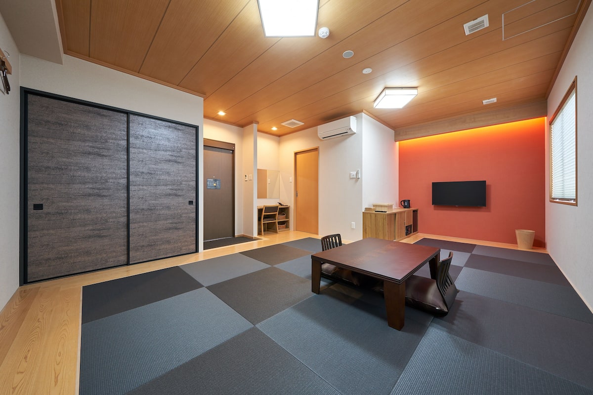 Ein Zimmer mit Tatami-Matten, einem Holztisch und einem Fernseher an der Wand.