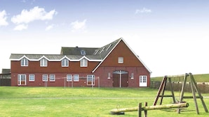 A red brick building with a gabled roof and a large central door, surrounded by a grassy field and a swing set. - Haus Seeblick (Neuwerk)