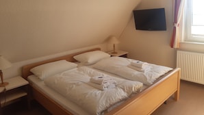 A neatly made bed in a room with a sloped ceiling, a bedside table with a lamp, and a television mounted on the wall. - Haus Seeblick (Neuwerk)
