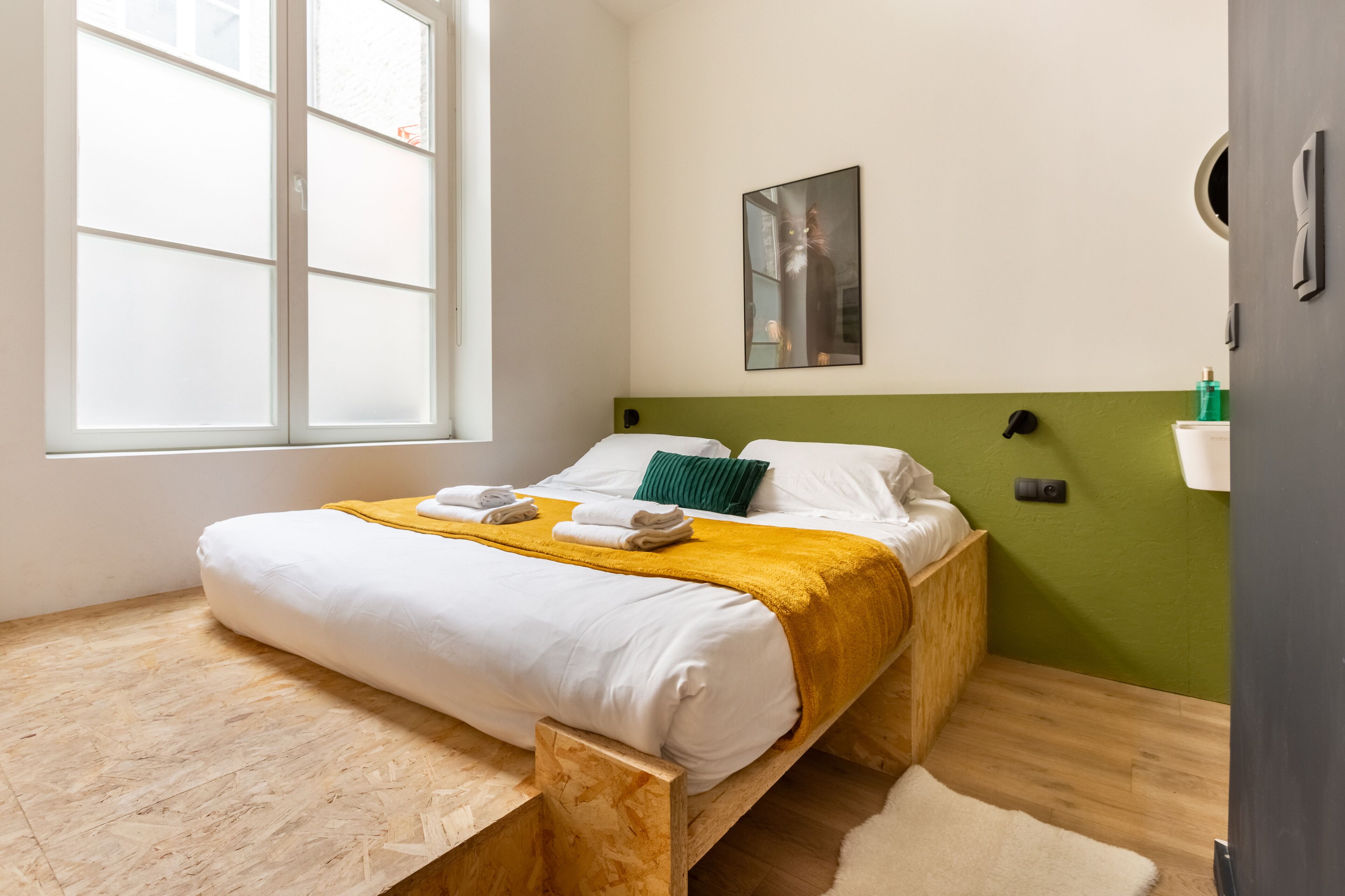 A neatly made bed with white linens and a yellow throw blanket, a green pillow, and a small bedside table with a lamp.