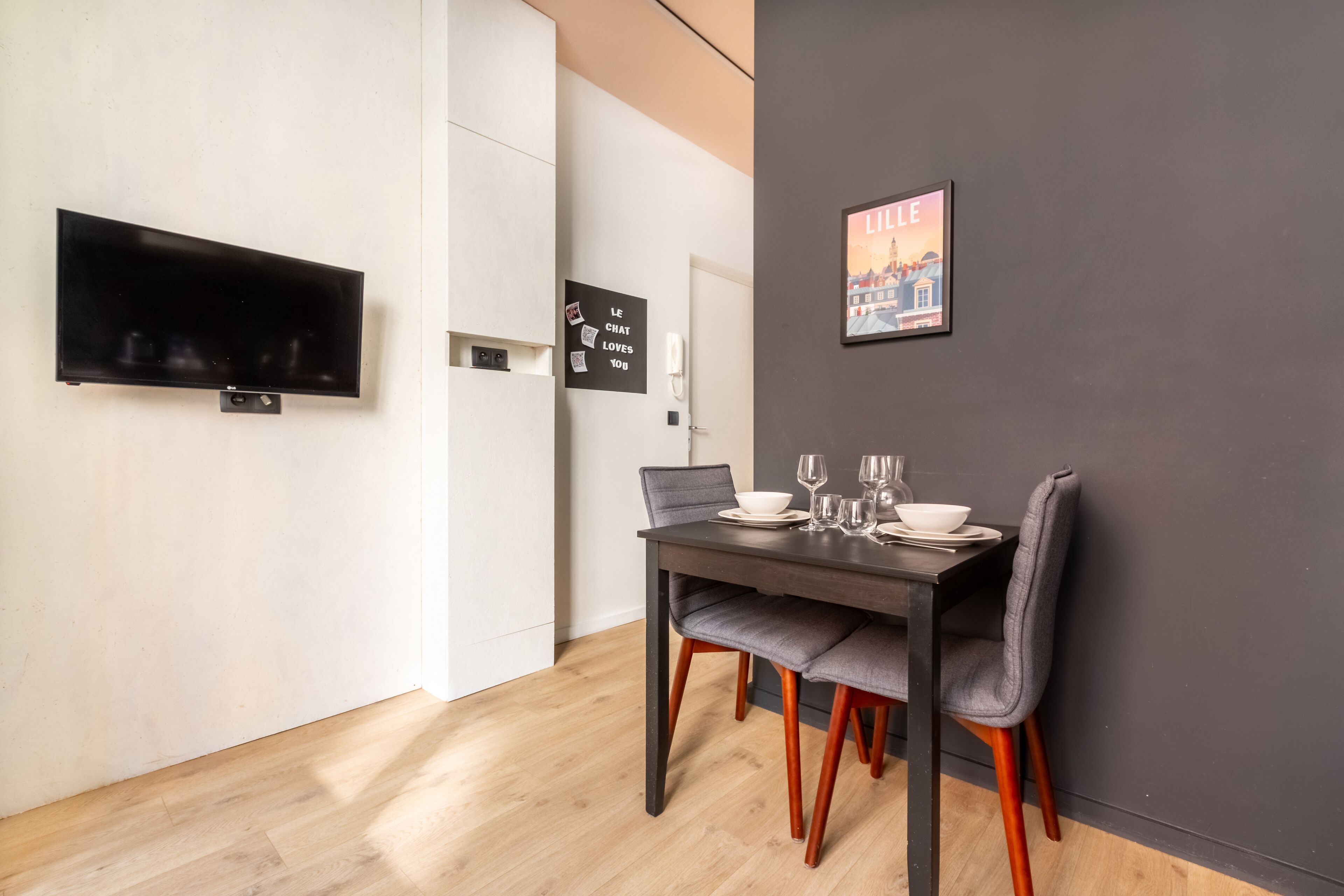 A modern dining area with a dark wooden table, grey upholstered chairs, and a flat-screen TV mounted on the wall.