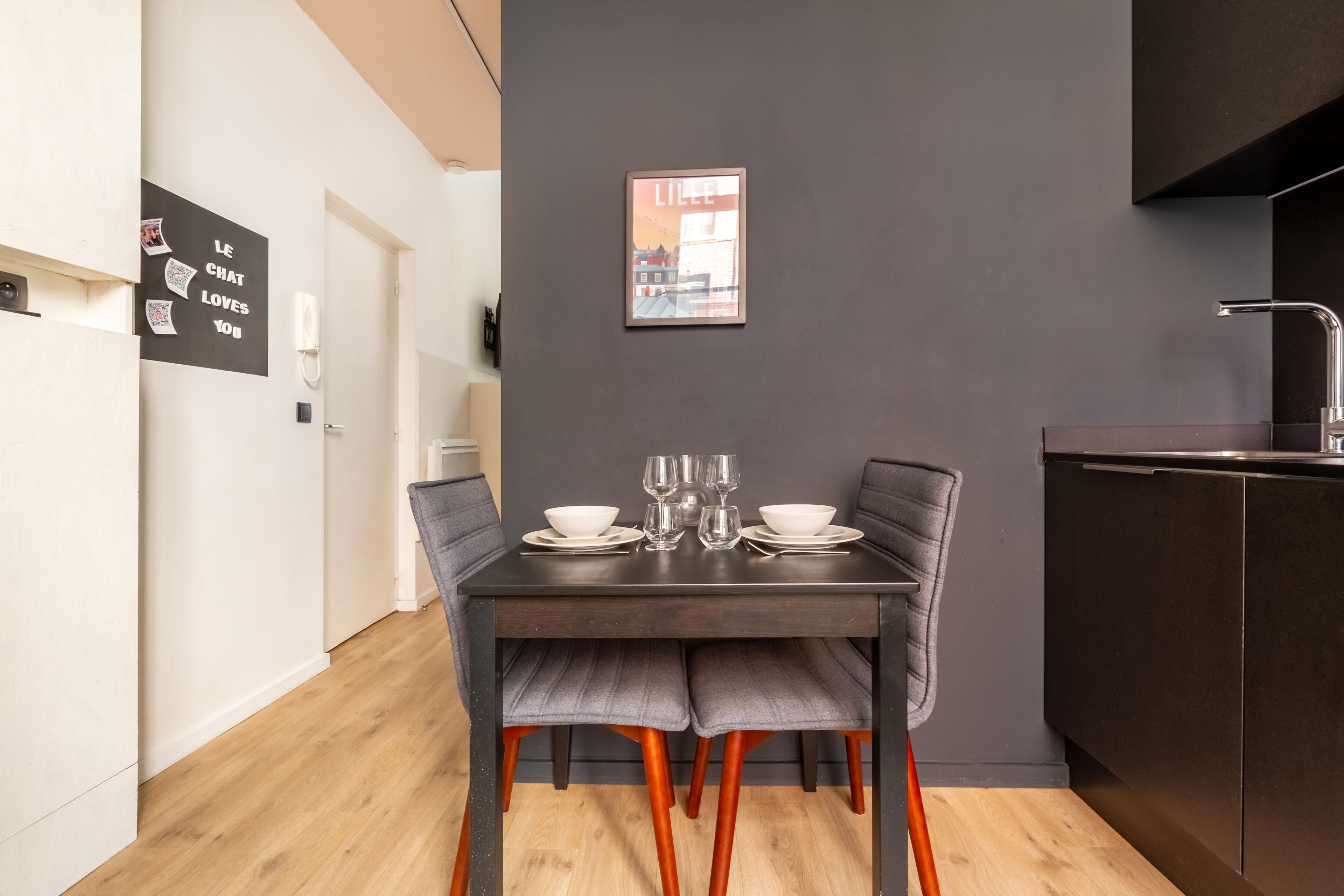 Un comedor moderno con una mesa de madera oscura dispuesta para dos, sillas grises con tapicería y un cuadro en la pared.