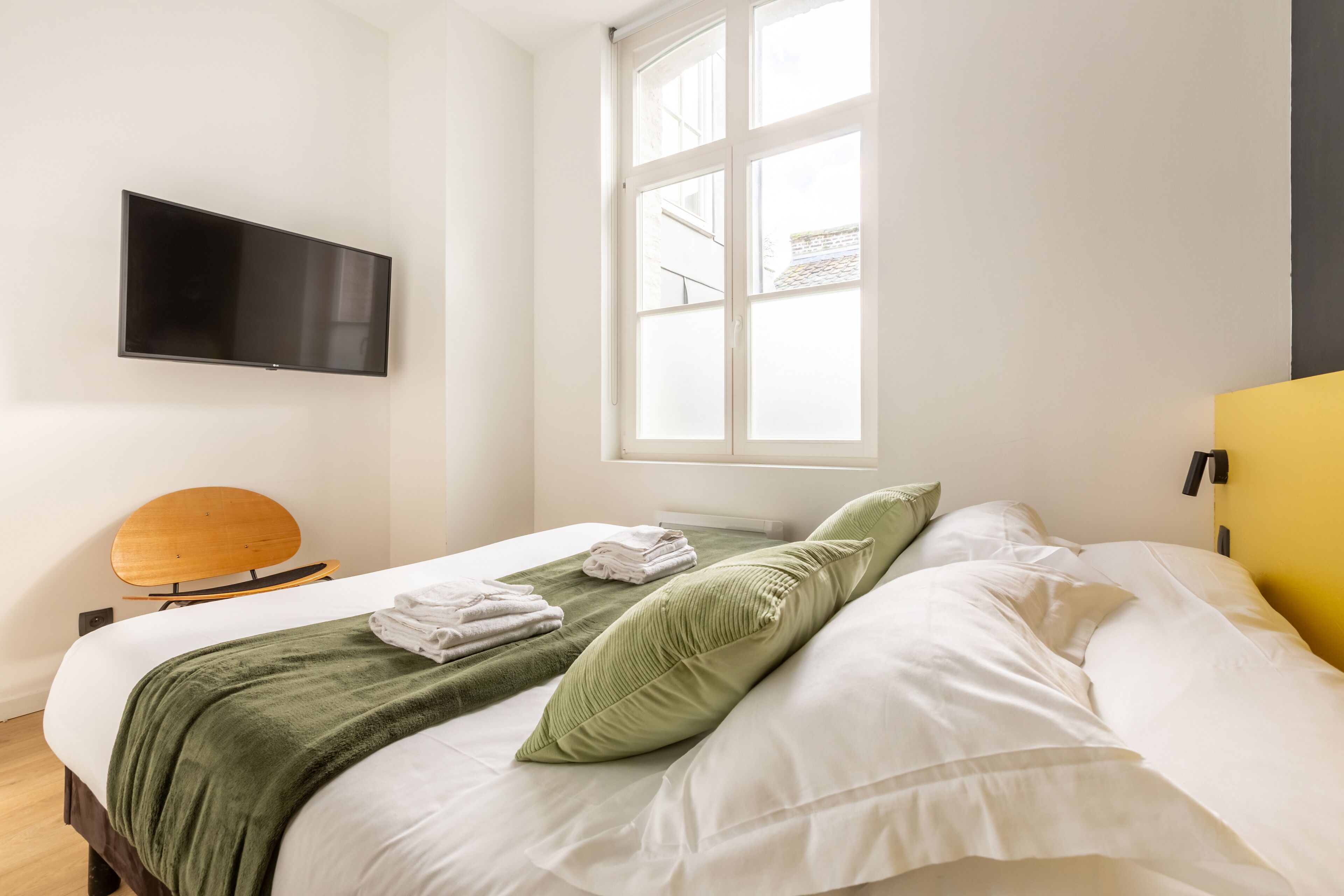 Una cama bien tendida con ropa de cama blanca y verde, un televisor de pantalla plana montado en la pared y una ventana con vistas a edificios.