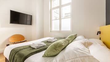 Una cama bien tendida con ropa de cama blanca y verde, un televisor de pantalla plana montado en la pared y una ventana con vistas a edificios.