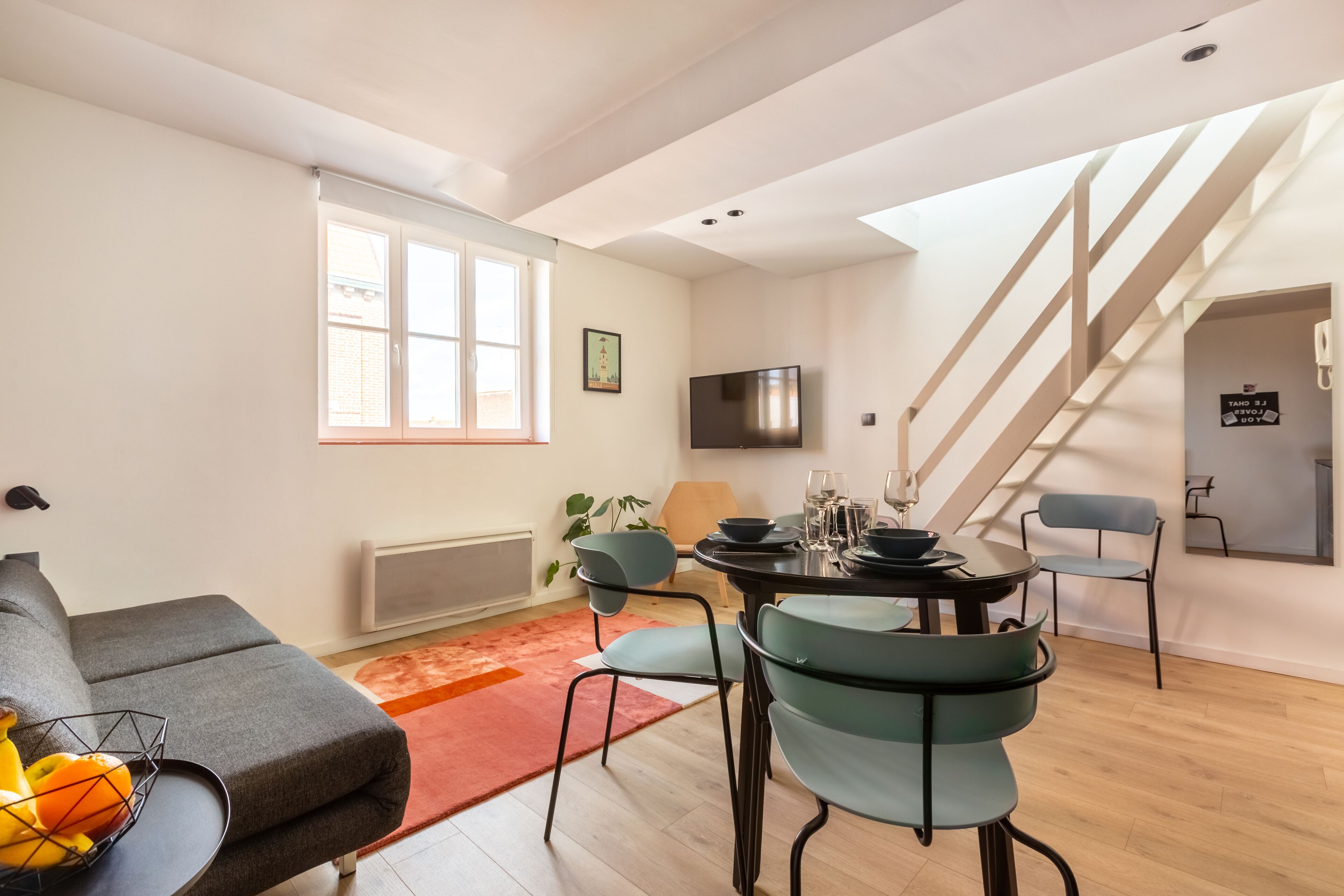 Un salon moderne avec un canapé, une table à manger et des chaises. On y trouve un escalier et un téléviseur fixé au mur.