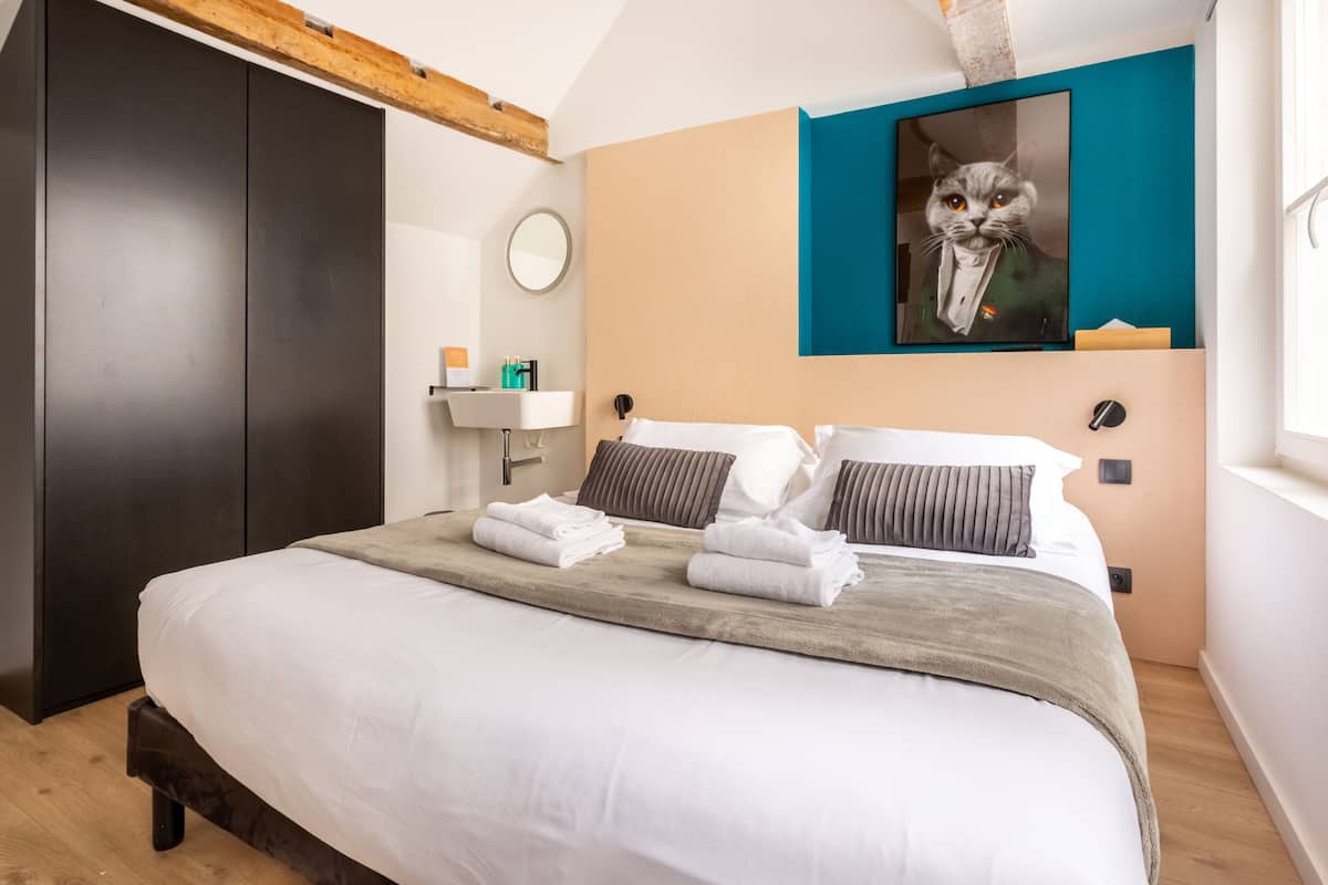 Un lit bien fait, agrémenté de serviettes, un miroir rond, un lavabo et un tableau représentant un chat accroché au mur.