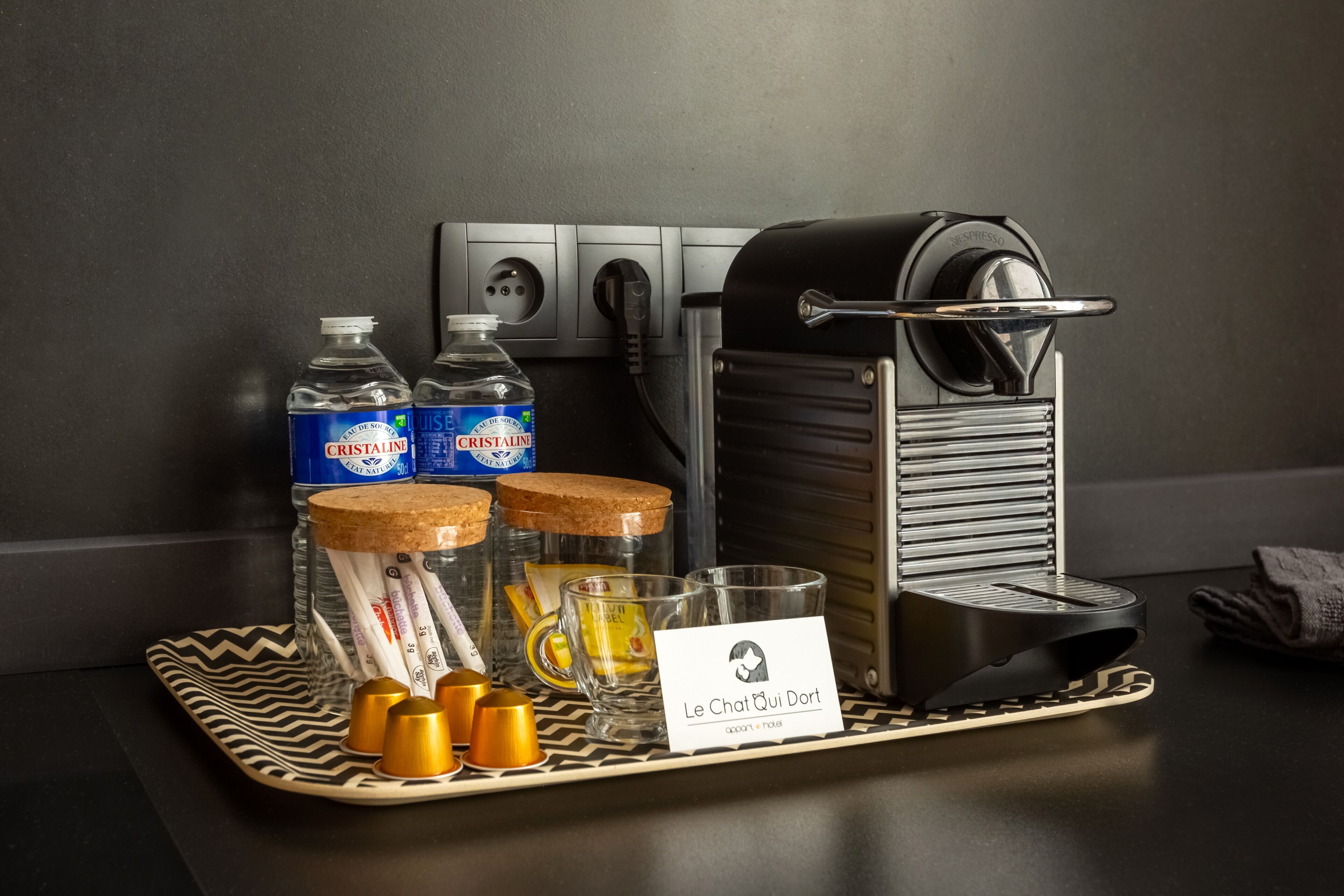 Una estación de café con una máquina de café, dos botellas de agua Cristaline, un vaso, y una bandeja con terrones de azúcar y una servilleta.
