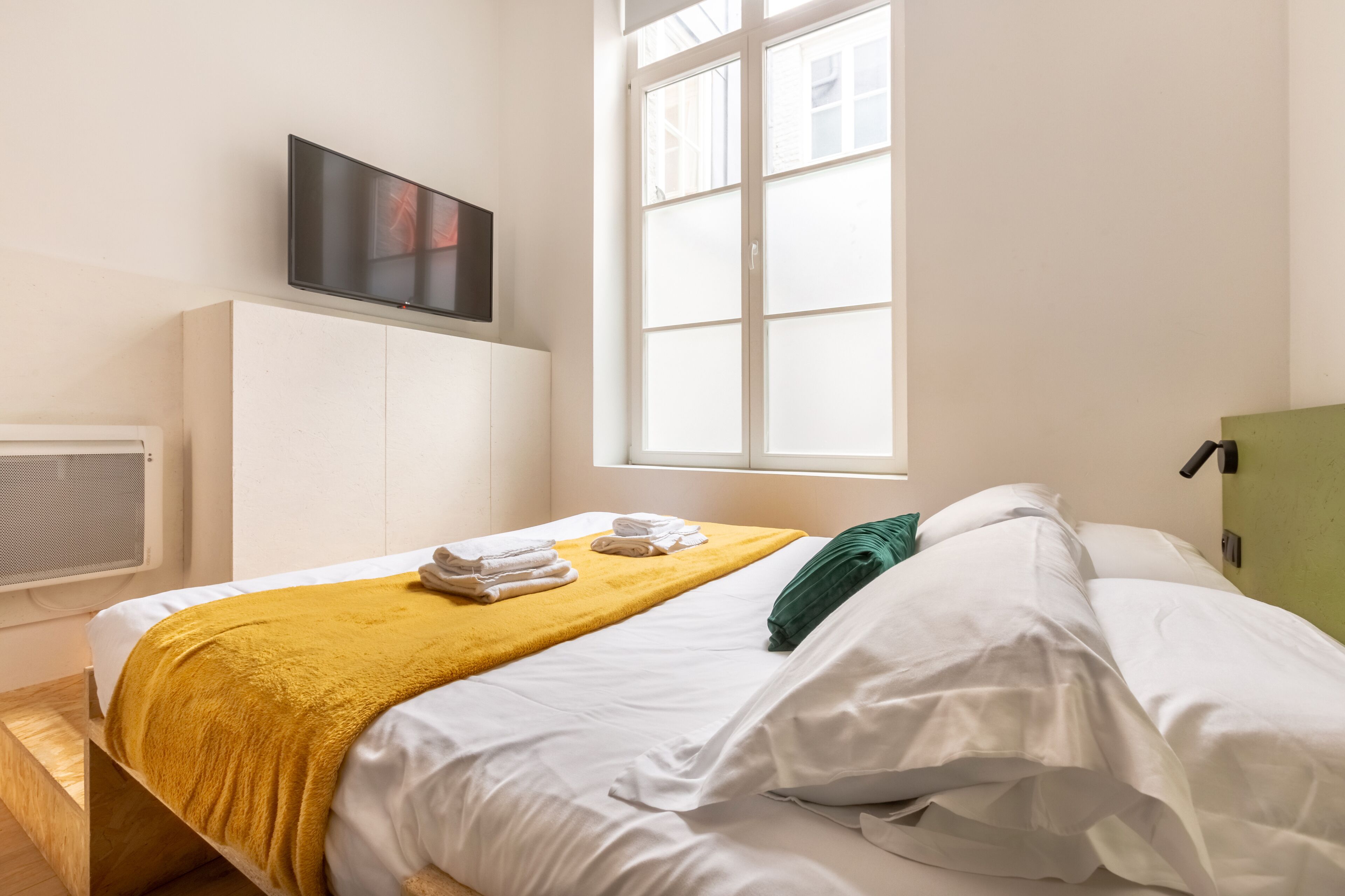 A neatly made bed with white linens and a mustard-colored throw, a flat-screen TV mounted on the wall, and a window with white blinds.